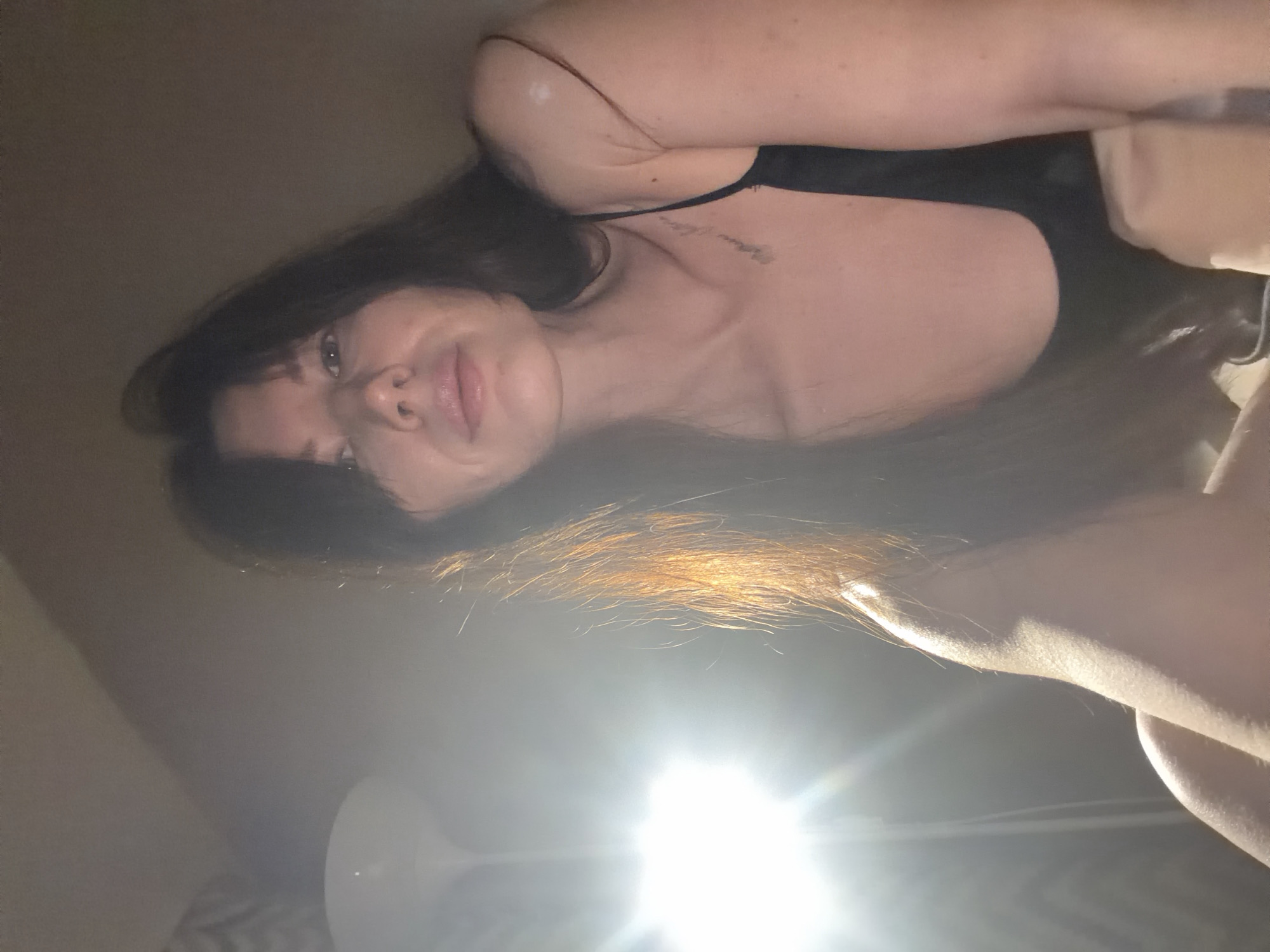A woman with long, dark hair is positioned looking downward at the camera. She is wearing a black tank top. A lamp is visible in the lower center of the frame, creating a bright flare. The location is not evident in the image.