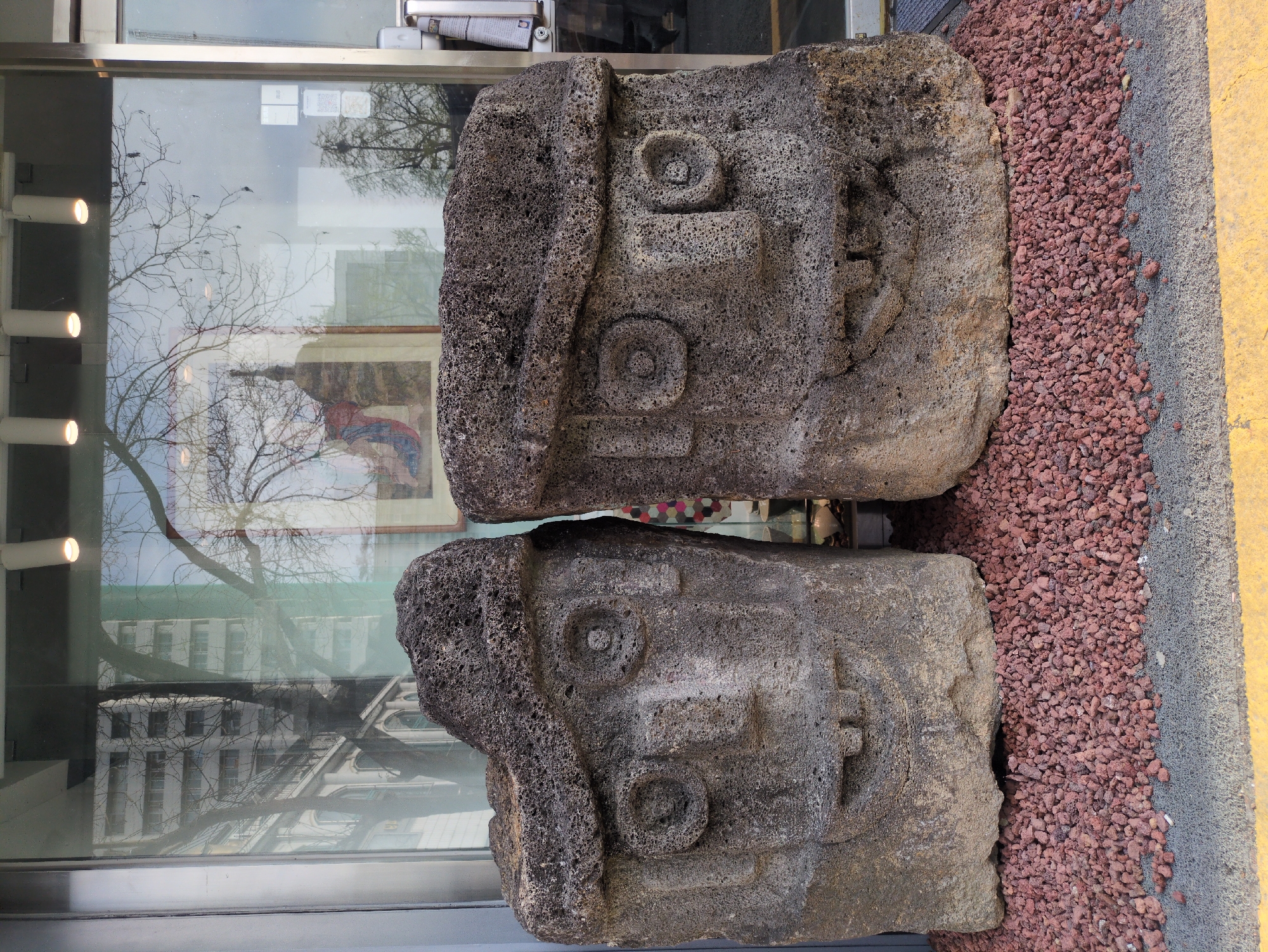 Two large, dark gray stone sculptures with carved faces are prominently displayed in front of a glass-fronted building. The sculptures have similar designs, featuring circular eyes, and are placed side-by-side on a bed of small, reddish-brown rocks. To the right, there is a gray curb with a yellow stripe. The building behind the sculptures has large windows reflecting trees and the surrounding cityscape. The location is Seoul, South Korea.