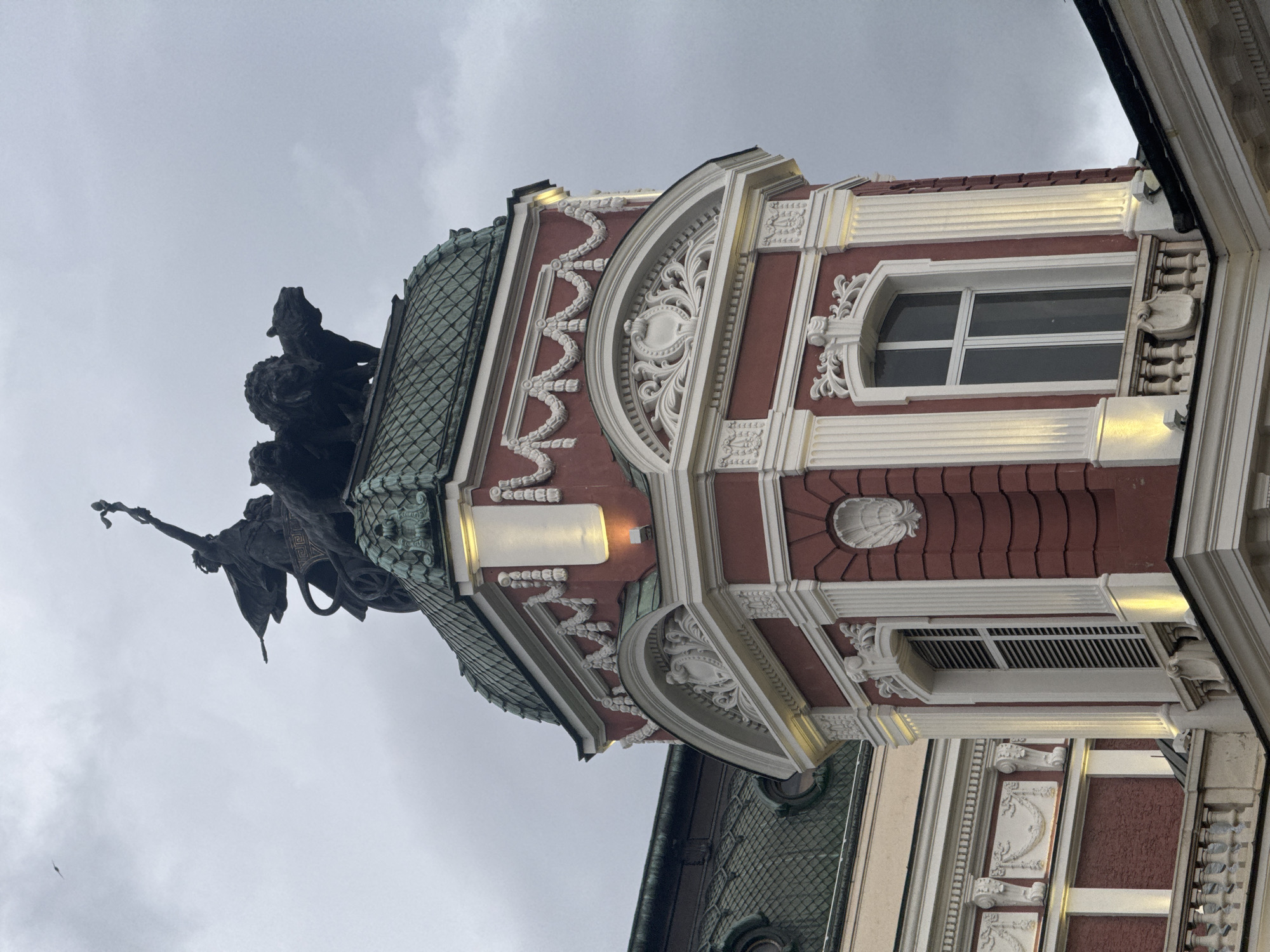 The image depicts a low-angle shot of the architecture and sculptural elements of a building in Sofia, Bulgaria. The architectural style features red brickwork accented by white decorative molding and trim. Windows with white frames are visible. The building is topped with a green, tiled roof. A statue of a man riding a lion is perched on top of the building. The sky is overcast.