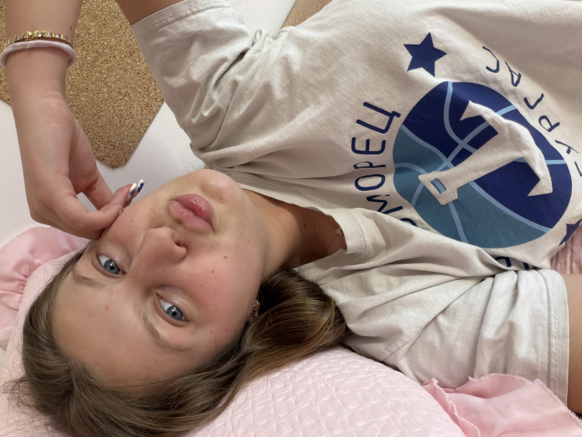 A young woman with light brown hair and striking blue eyes lies relaxed, looking directly at the camera. Her head is tilted, and her expression is neutral with slightly parted, full lips. She is wearing a light beige or off-white short-sleeved t-shirt. On her right wrist, she wears a golden or jeweled bracelet over a light pink fabric band. Her fingernails on the visible right hand are painted with a distinctive white and blue design.
The t-shirt features a prominent blue graphic with a white anchor inside a globe-like design, and a dark blue star above it. Several Cyrillic letters are visible on the shirt, including "МОРЕЦ" (Morets, meaning "sailor" or "seaman") and parts of "КУРОРТ" (Kurort, meaning "resort"), suggesting a maritime or resort theme consistent with the location context of Burgas, Bulgaria.
She is resting on a pink quilted pillow, and her hair is spread out behind her. The background includes a light, possibly white, surface and a textured cork board material. The scene takes place indoors, likely in a bedroom or a comfortable living space.