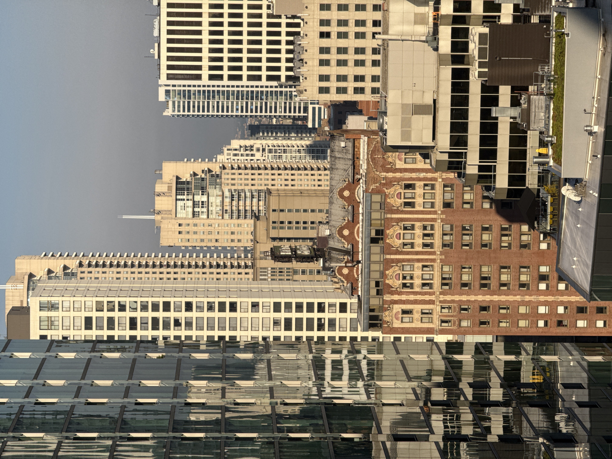 This media file displays a vertical, close-up view of several tall buildings in an urban setting, likely Chicago, USA. The scene is dominated by the architectural features of the buildings, which exhibit a range of styles. The top portion of the image features a pale building with rows of dark rectangular windows. Below this, a complex of buildings with cream-colored facades and numerous evenly spaced windows stretches downwards. Further down, a distinctive brick building with ornate architectural details and smaller windows is visible. The bottom of the image is filled with the reflective glass facade of a modern skyscraper, mirroring the sky and other buildings.
The image appears to have been taken during the day, with clear, bright lighting. The sky is a pale, hazy blue, suggesting a possibly overcast or smoggy day, common in urban environments. There is no discernable activity taking place within the visible parts of the buildings, and no people are clearly visible. The focus is entirely on the structures themselves and their interaction with light.
Notable details include the sharp lines of modern architecture juxtaposed with the more traditional brickwork. The reflections in the glass building at the bottom create a complex interplay of light and form, adding depth and visual interest to the scene. The overall impression is one of a dense urban landscape, characterized by towering structures and a somewhat muted color palette, with the exception of the warm tones of the brick building. There is no visible text within the image.