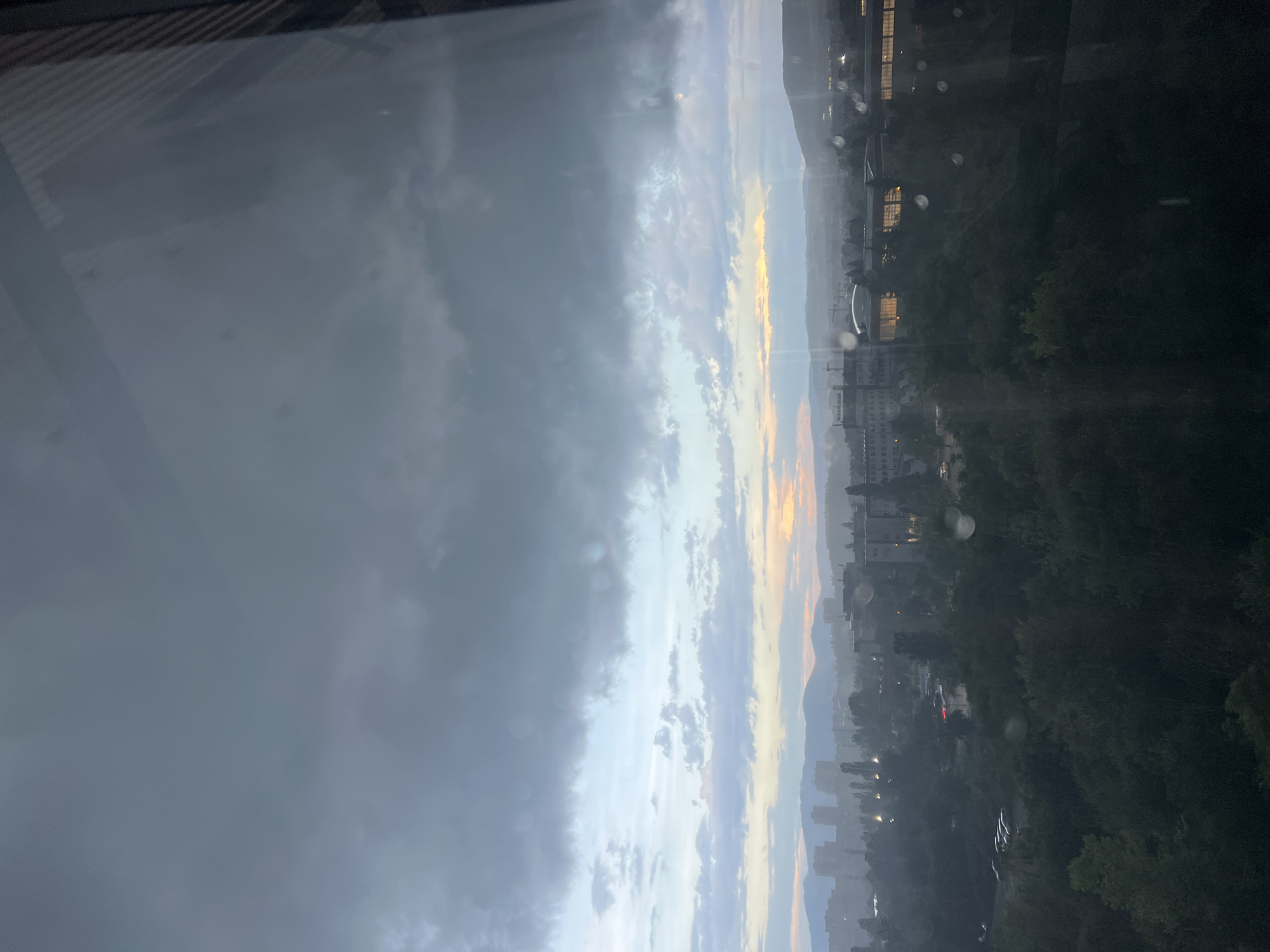 The image captures an expansive view of a cityscape and natural landscape, likely from an elevated perspective, possibly through a window with reflections and droplets. The sky dominates the upper portion, featuring a dramatic contrast: heavy, dark grey clouds are prominent on the left, while the right displays lighter clouds illuminated by warm orange and yellow hues, suggesting either sunrise or sunset.
Below the horizon, distant mountains or hills are visible, partially obscured by atmospheric haze. Closer to the foreground, an urban environment unfolds with numerous buildings, some appearing residential or commercial, and a few lights are visible in the windows or on the streets, implying it is either dusk or dawn. A significant portion of the mid-ground and foreground is covered by dark green foliage, indicative of parks or forested areas within the city. One crop shows a glimpse of what might be a street with parked cars and a distant red light.
No specific human activity is discernible. The overall impression is one of a transitional time of day with varying weather conditions. The scene is consistent with a city like Sofia, Bulgaria, nestled against a mountainous backdrop. No legible text is visible in the image.