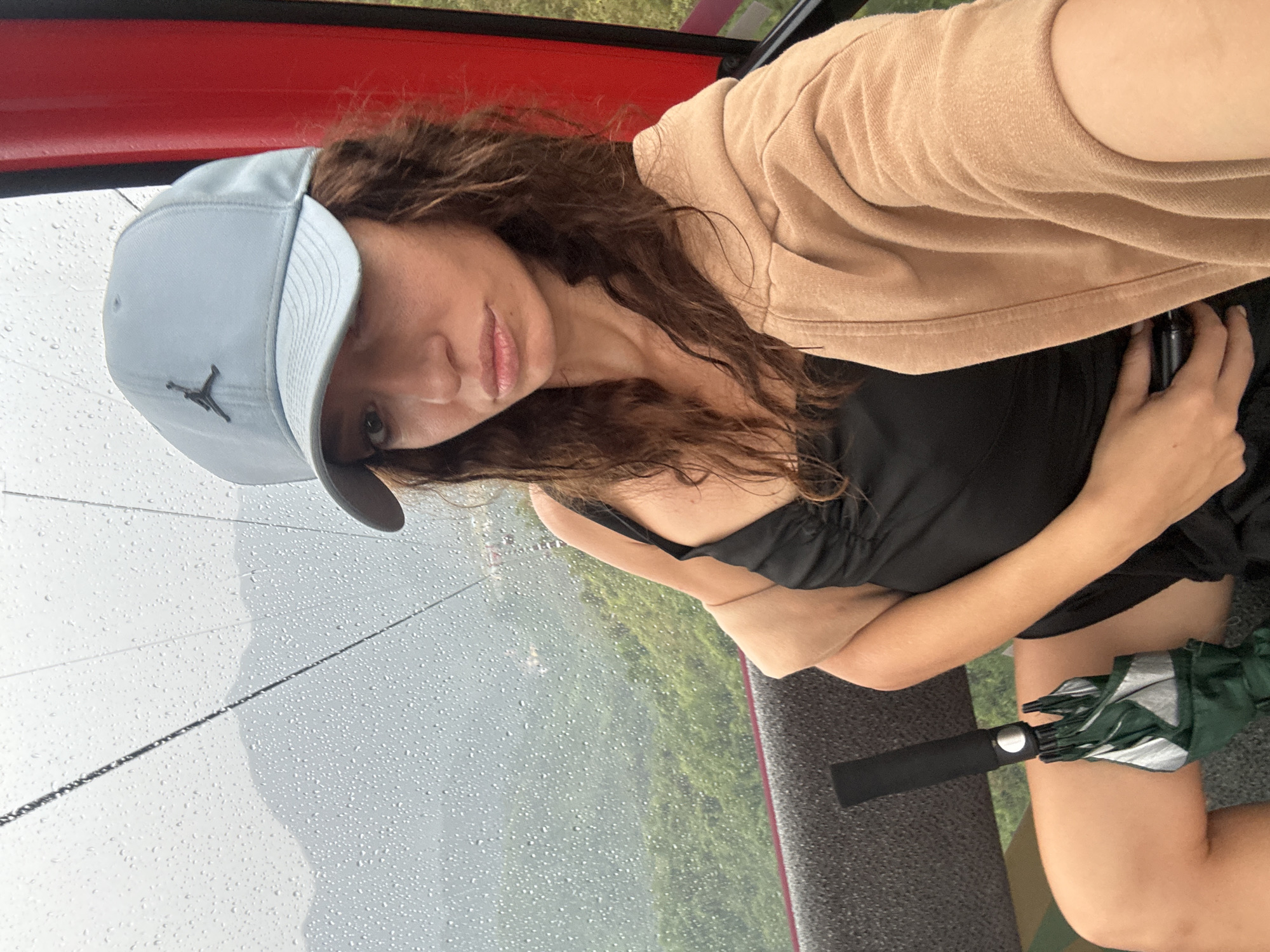 The image features a woman taking a selfie inside what appears to be a cable car cabin, with a rainy, misty landscape visible through the window. The woman, with long wavy brown hair, looks directly at the camera with a calm, neutral expression. She is wearing a light blue baseball cap featuring a dark "Jordan" (Jumpman) logo on the front. Her attire includes a black top, layered with a beige or tan long-sleeved top or light jacket. Her right hand is near her chest, holding a dark object, possibly a phone. A folded dark green and silver umbrella rests on her lap, indicating the wet weather.
The setting is an enclosed space, likely a cable car cabin, with a prominent red frame visible at the top and side. The window is covered in numerous raindrops, blurring the exterior view. Beyond the rain-streaked glass, indistinct green trees and mountainous terrain can be seen, partially obscured by the weather conditions, suggesting an overcast daytime. The scene conveys a sense of travel or exploration in a scenic area. Given the context, the location is Đà Nẵng, Vietnam.