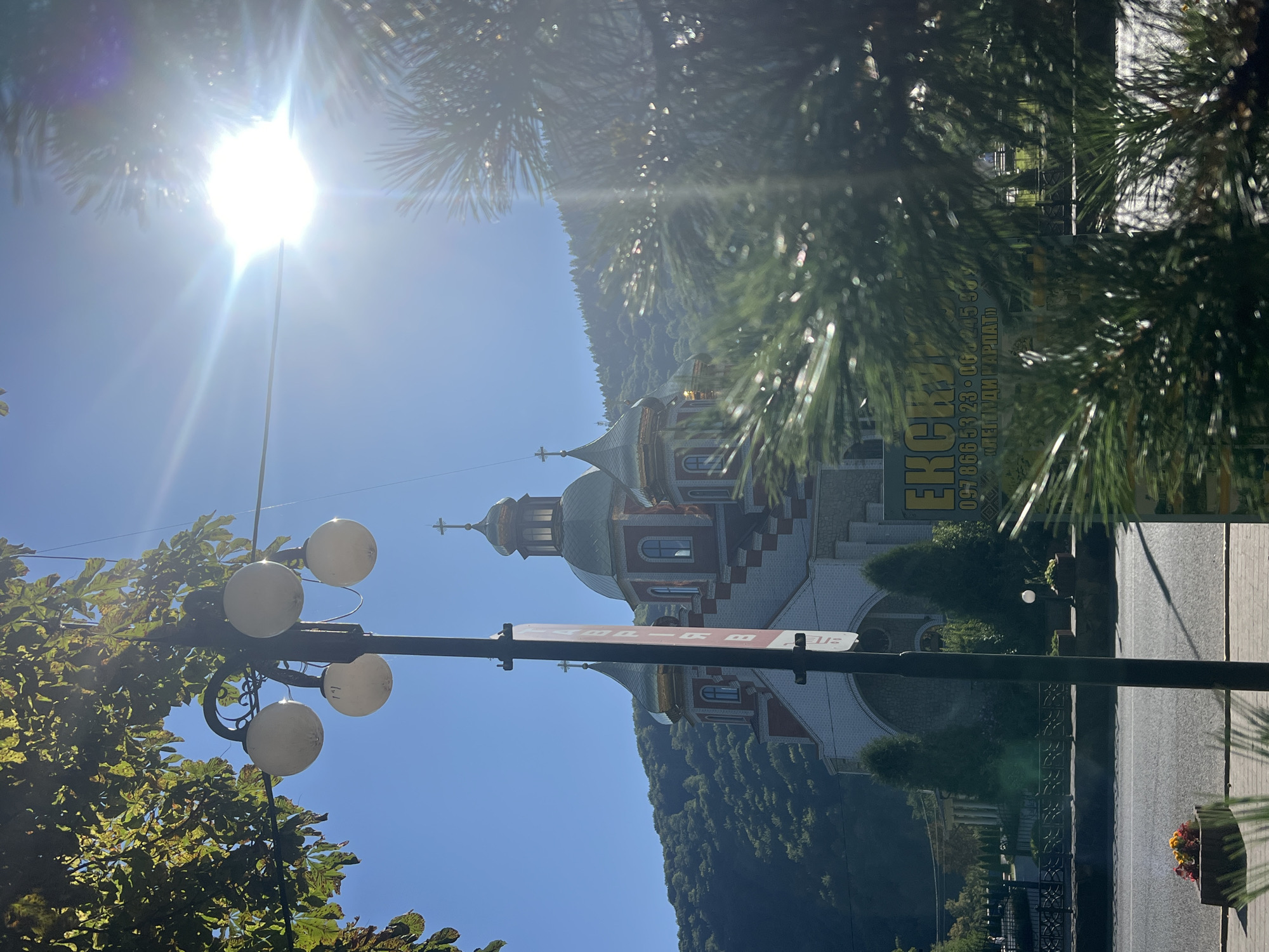 In Yaremche, Ukraine, a street scene unfolds under a bright, sun-drenched sky with a noticeable sun flare. A tall lamppost with four spherical lights stands prominently in the foreground, partially obscuring a red and white church with green-domed towers and crosses. The church is nestled against a backdrop of lush, green, tree-covered hills. In front of the church, several small, conical evergreen trees are visible, and a street with a sidewalk runs across the lower part of the frame. A planter with colorful flowers sits on the sidewalk. In the middle ground, a green sign with yellow text that reads "ЕКСКЛЮЗИВ" is partially visible, along with a phone number and "ЛЕГЕНДИ КАРПАТ". The foreground is blurred by the needles of a pine tree.