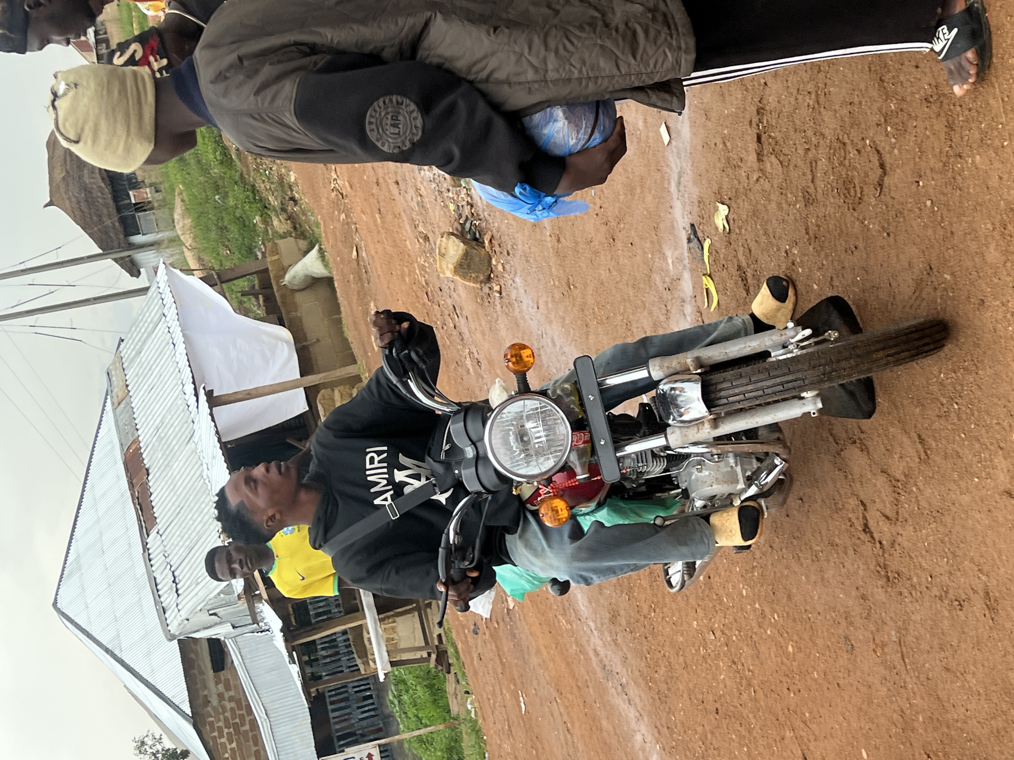 In Durumi, Nigeria, a young man sits astride a motorcycle, his legs straddling the machine. He wears a black hoodie with the word "AMIRI" emblazoned across the chest in white. His feet are clad in light brown sandals. He is looking forward, his expression difficult to discern.
Behind him, another young man in a bright yellow shirt with a Nike logo is visible. Further back, a person wearing a brown jacket and a light-colored head covering is seen holding a blue plastic bag. The scene appears to be outdoors, with a dirt path or road underfoot. The background shows a collection of structures, some with corrugated metal roofs and others with thatched roofs, suggesting a rural or semi-urban environment. Some debris is scattered on the ground. The overall lighting suggests it is daytime. The mood seems casual, with people gathered around the motorcycle.