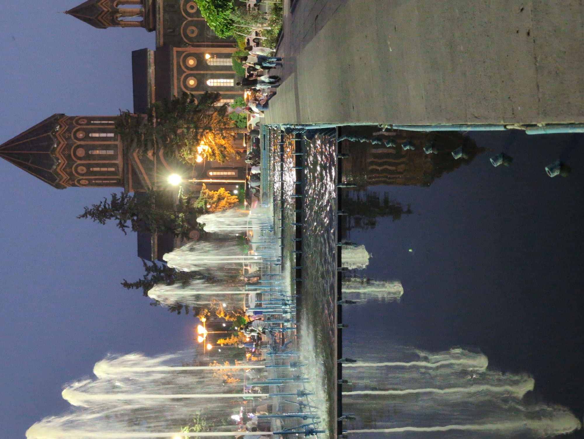 The image depicts a fountain in front of a large, ornate building that appears to be a church, possibly St. Mary's Cathedral in Gyumri, Armenia, given its architectural style. The scene is illuminated by streetlights and the glowing lights from the building, creating a twilight atmosphere. Several jets of water from the fountain are illuminated, casting reflections in the dark water. A paved walkway runs alongside the fountain, and several people are visible walking along it and gathered near the fountain's edge. The building behind the fountain is characterized by its domed roofs and arched windows. Trees are also present, adding to the park-like setting.