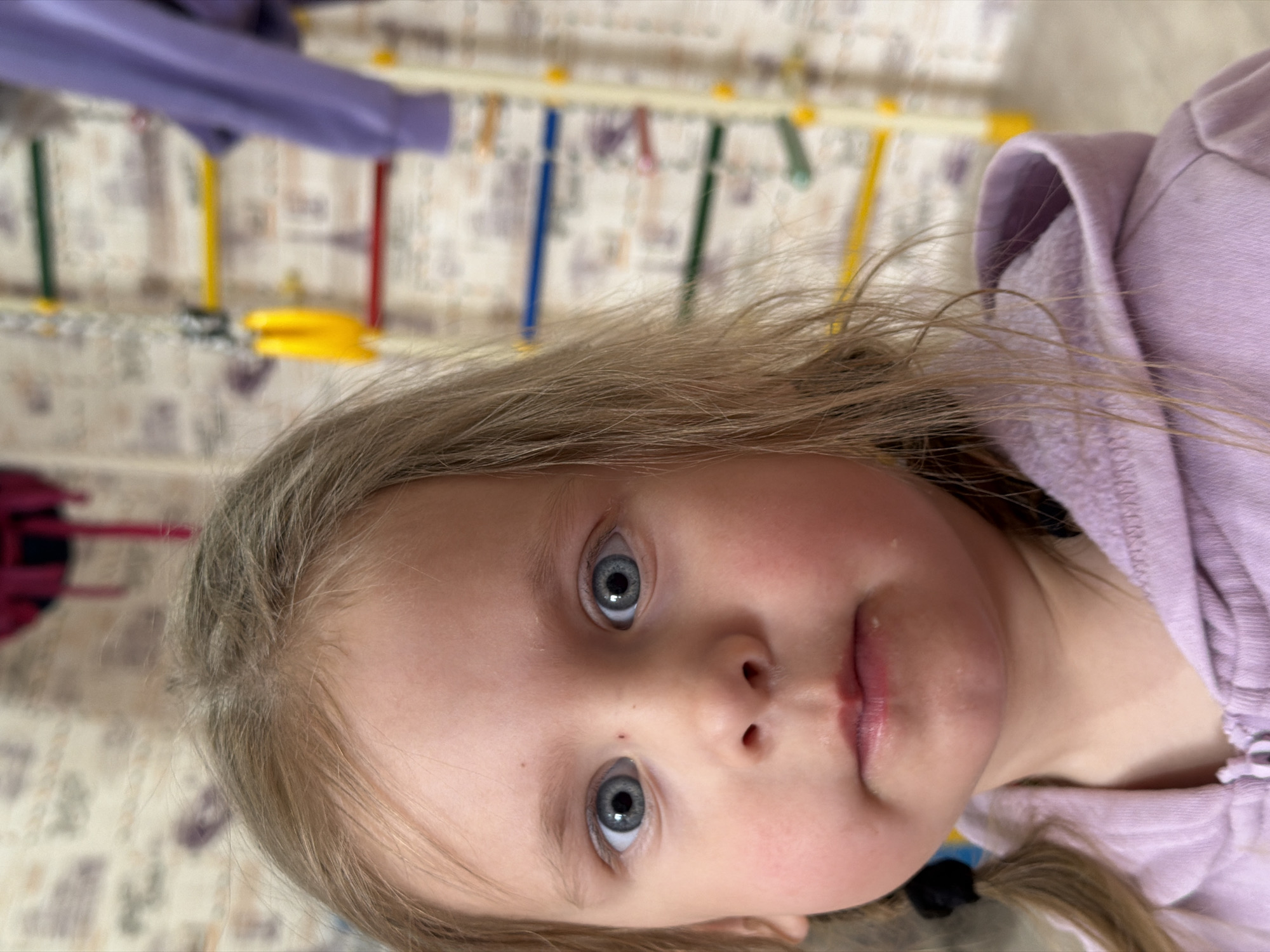 A close-up, slightly rotated view shows a young girl with light blonde hair and striking blue eyes. She wears a light purple hooded sweatshirt. The background features wallpaper with a repeating pattern and a colorful wall-mounted climbing structure with rungs of yellow, blue, green, and red. Although the city is mentioned, there is no way to verify the location from this image.