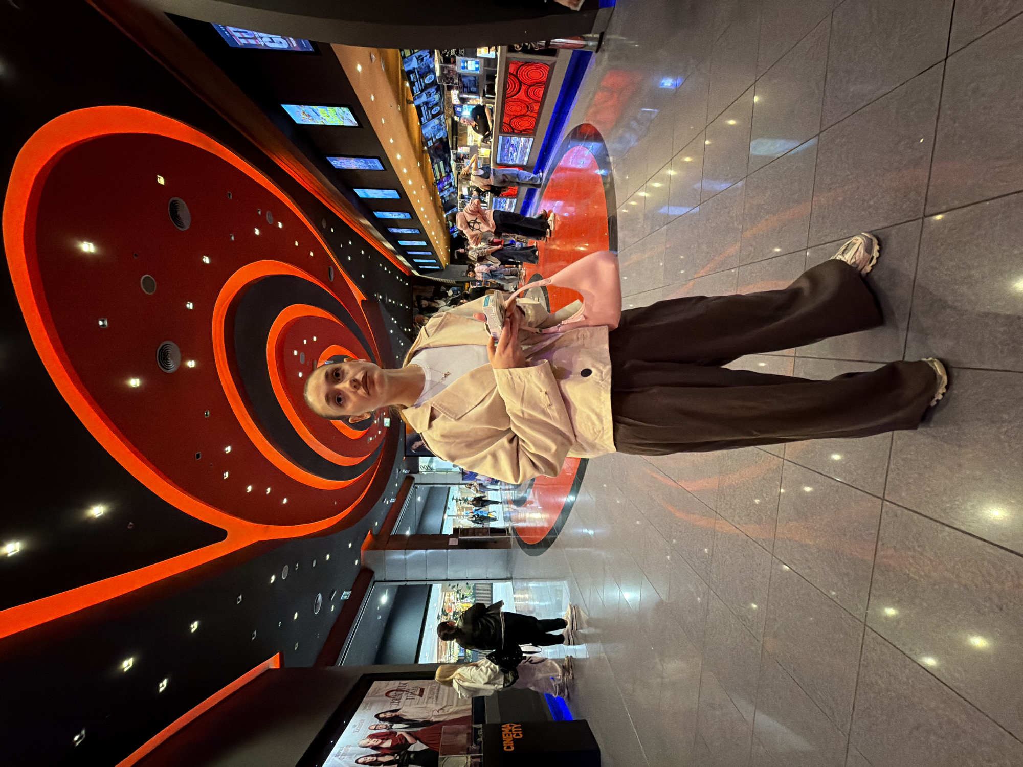 The image is taken inside a Cinema City location, likely in Sofia, Bulgaria, based on the "Cinema City" signage visible at the bottom of the frame.
A young, fair-skinned individual is centered in the foreground, facing the camera. They are wearing a light-colored jacket, a white top, and dark trousers. They are holding what appears to be a beverage and a light pink handbag.
The background features the interior of the cinema complex, with a long corridor extending into the distance. The corridor has a distinctive ceiling design with illuminated orange and black concentric circles. Several people are walking along the corridor. On the left side of the corridor, screens display movie information. The flooring is tiled with a reflective surface. A movie poster stand is partially visible in the lower left corner.