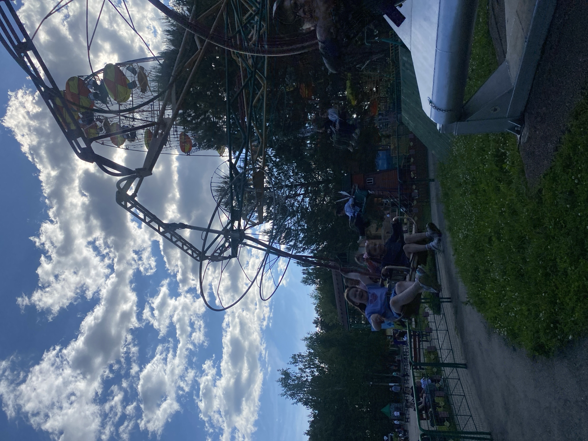 This image depicts an amusement park in Chaikovsky, Russia, under a partly cloudy sky. The scene features two operational amusement rides and numerous park visitors.
In the foreground and mid-ground, a multi-seat swing ride, characterized by a metallic framework, is active. Two young females, likely children, are seated in individual swings in the immediate foreground; one wears a light blue top and shorts, while the other wears a darker top. Both exhibit open-mouthed expressions. Additional figures are visible in other swings on the ride further back.
To the right in the background, an operational Ferris wheel is prominent. It features multiple gondolas, distinctly colored in yellow and orange/red patterns. Occupants are discernible within some of these gondolas. The sun, partially obscured by clouds, casts backlighting on the ride structures, creating areas of silhouette.
The environment includes dense green tree cover, composed of both coniferous and deciduous species, forming a natural backdrop. A paved pathway is visible to the left, running alongside a green metal fence. The ground area in the foreground consists of green grass interspersed with small white flowers. In the bottom-right corner, a metallic ground support structure is partially visible, featuring a chain and an electrical grounding symbol.
The sky is blue with scattered white and grey cumulus clouds. Numerous indistinct figures, including adults and children, are distributed throughout the background areas of the park, indicating general recreational activity. The overall lighting is natural daylight, suggesting either late morning or afternoon conditions.