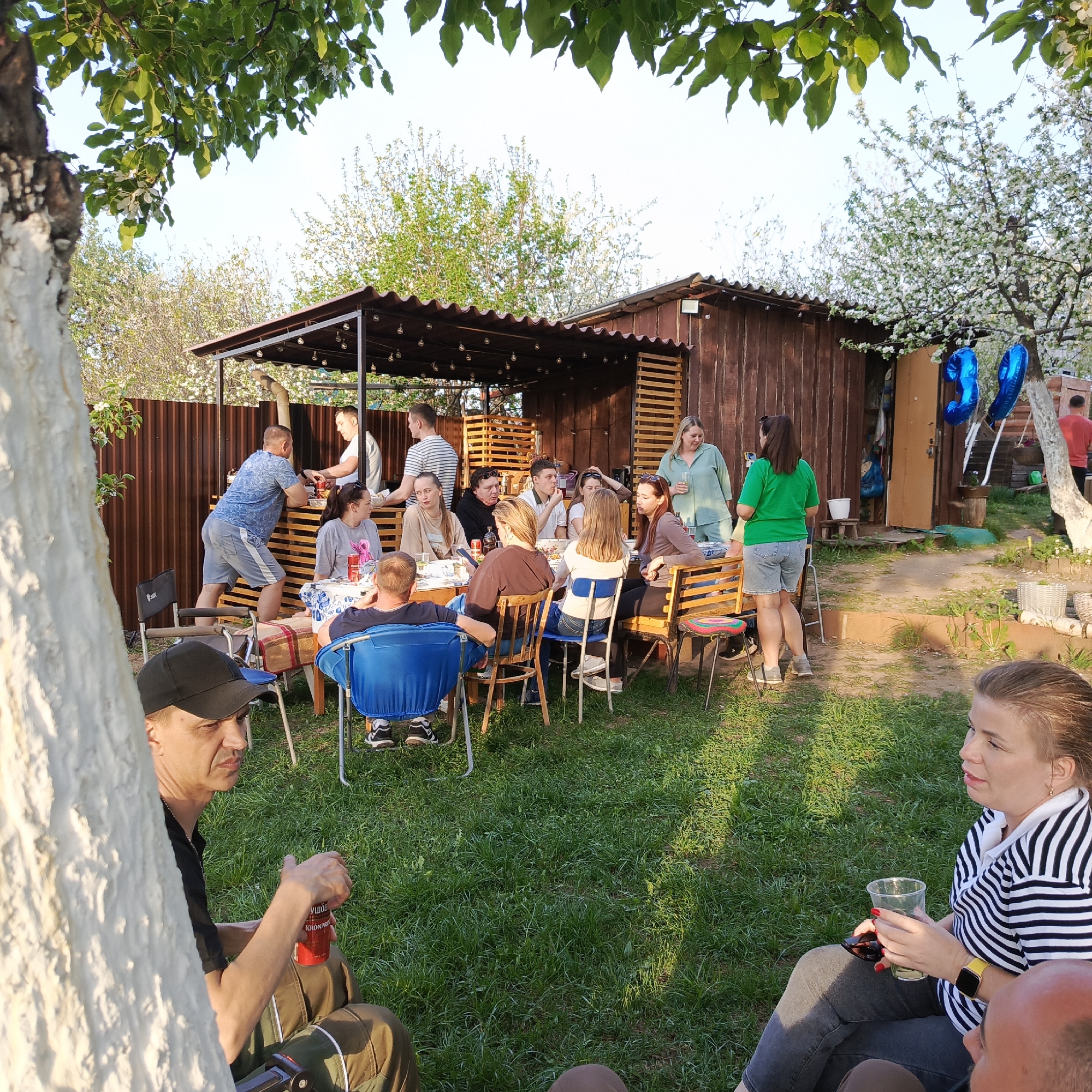 The image depicts a group of people gathered outdoors, likely in a backyard in the city of Chaikovsky, Russia. A large table is surrounded by people sitting on chairs and benches, suggesting a meal or gathering. Some individuals are standing, engaging in conversation. A structure with a roof and lights hangs overhead, possibly a pergola or outdoor bar area. A wooden shed is visible in the background, decorated with blue balloons. In the foreground, two people are sitting and conversing, with one holding a clear plastic cup. There is a tree in the foreground of the image. The scene suggests a social event or celebration.
