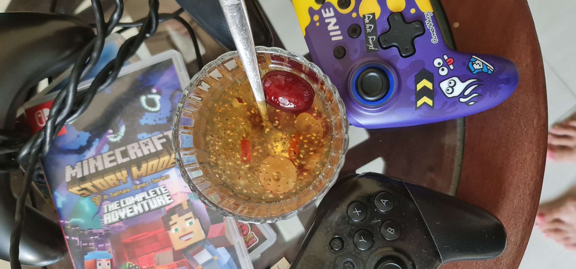 The image captures an indoor scene, likely a living space, with various objects arranged on a surface. A glass bowl filled with a light-colored liquid containing seeds, fruit slices, and a large red fruit sits at the center. A spoon rests inside the bowl. To the left of the bowl is a Nintendo Switch game case titled "Minecraft Story Mode: The Complete Adventure." Two game controllers, one black and one purple with yellow accents and cartoonish designs, are positioned to the right and below the bowl, respectively. Black cords snake around the objects. The surface appears to be a brown, possibly wooden table. In the lower right corner, a pair of feet with pink nail polish are partially visible. There is no explicit evidence within the image that indicates it was taken in the city of Da Nang, Vietnam.