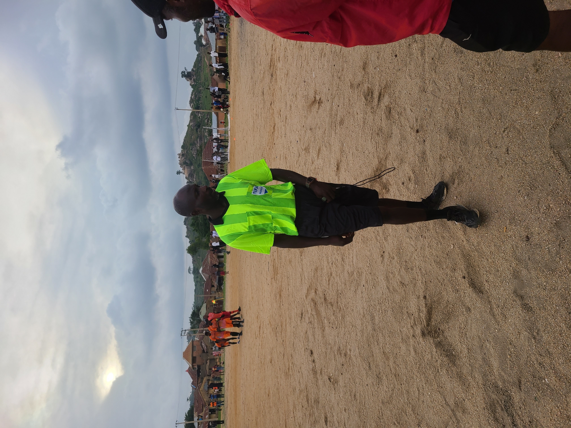 A soccer game is taking place in the city of Unknown, Unknown. The field is sand. A soccer referee wearing a bright green vest is on the field. A group of people are wearing orange soccer uniforms. There are several people watching the game. There are some buildings in the background.