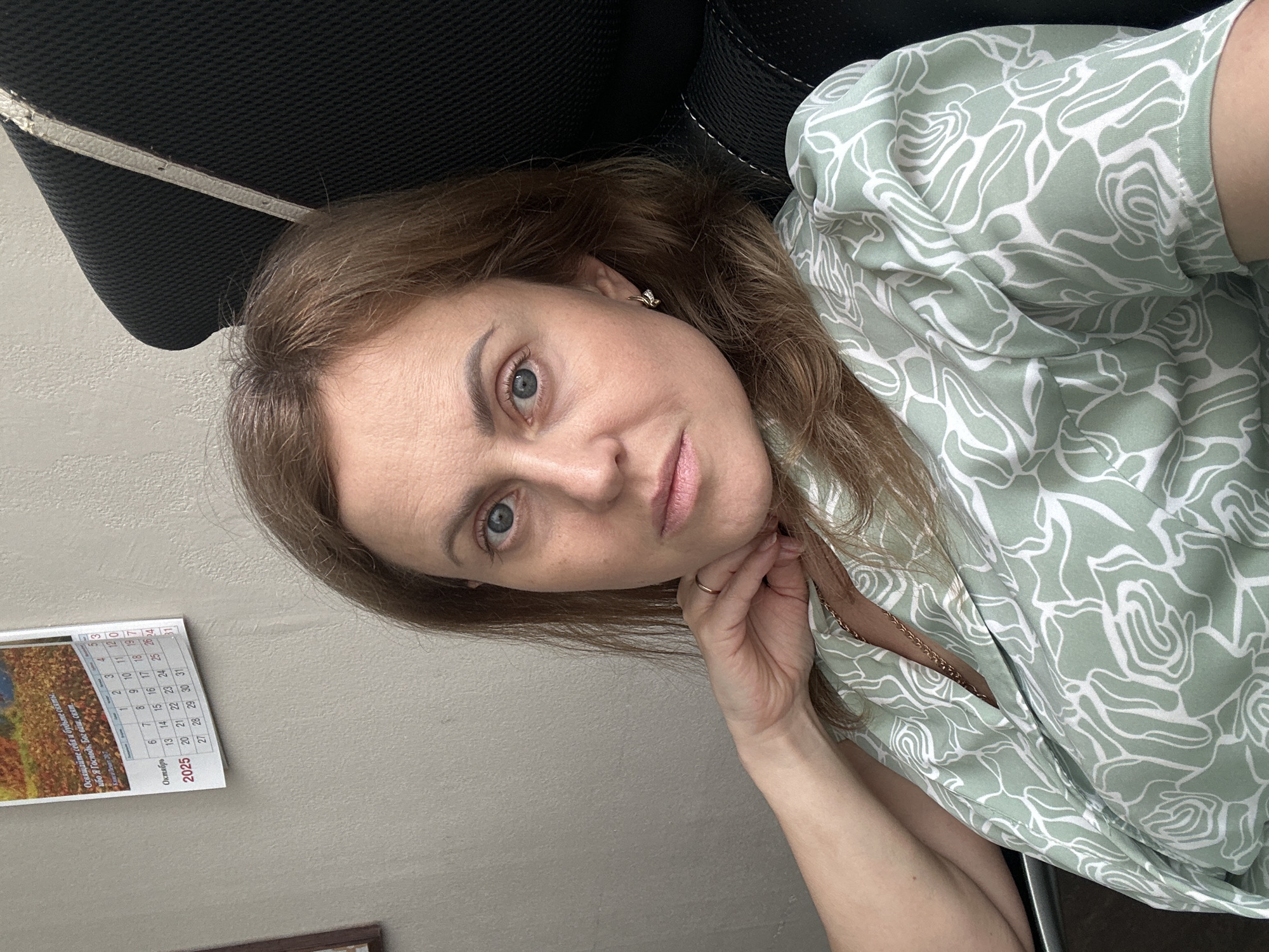 A woman with shoulder-length brown hair and blue eyes is captured in a close-up, head-and-shoulders shot. She is seated, with her left arm resting on her side and her hand touching her neck. She is wearing a light green blouse with a white floral pattern and a gold chain necklace. Her gaze is directed towards the camera. In the background, to the left, a calendar for October 2025 is visible. The wall behind her is a neutral, textured gray. To the right, the dark mesh material of a chair is present. No specific location within Sochi, Russia, can be determined from the image.