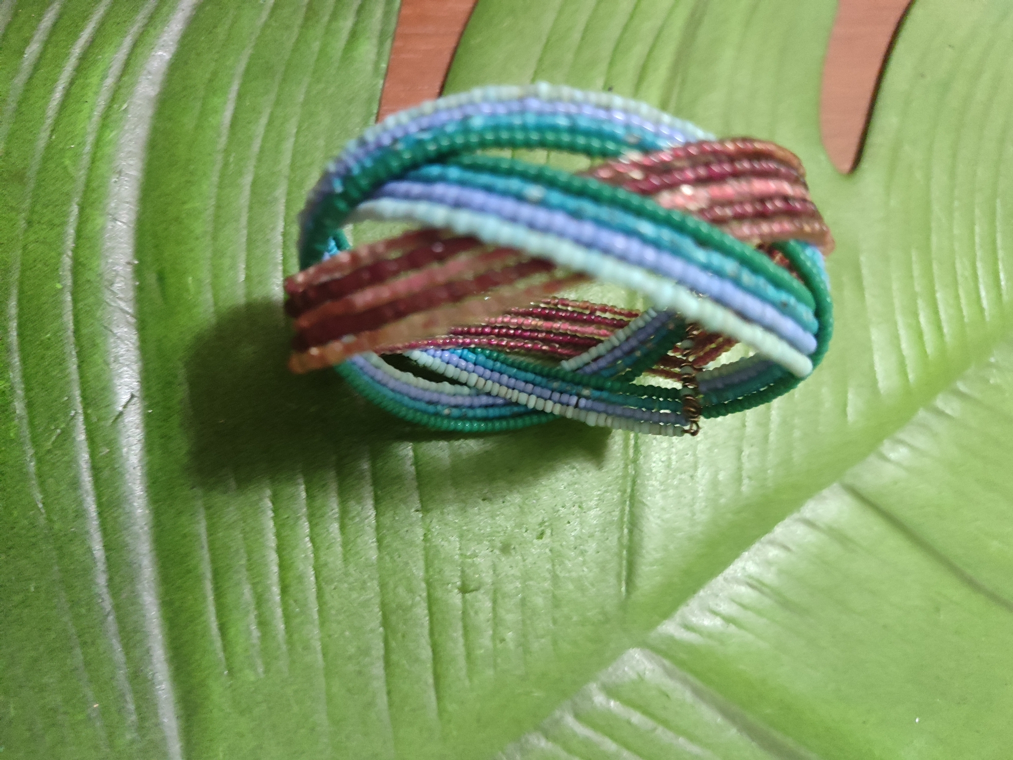 A beaded bracelet with multiple strands of turquoise, purple, and pink beads is displayed on a large, textured green leaf. The bracelet is coiled and interwoven, with some strands appearing more tightly wound than others. The leaf has prominent parallel veins. The background is out of focus, showing hints of a wooden surface. The image is a close-up shot with shallow depth of field. The location is not specified in the image, and therefore cannot be identified as Mykolaiv, Ukraine.