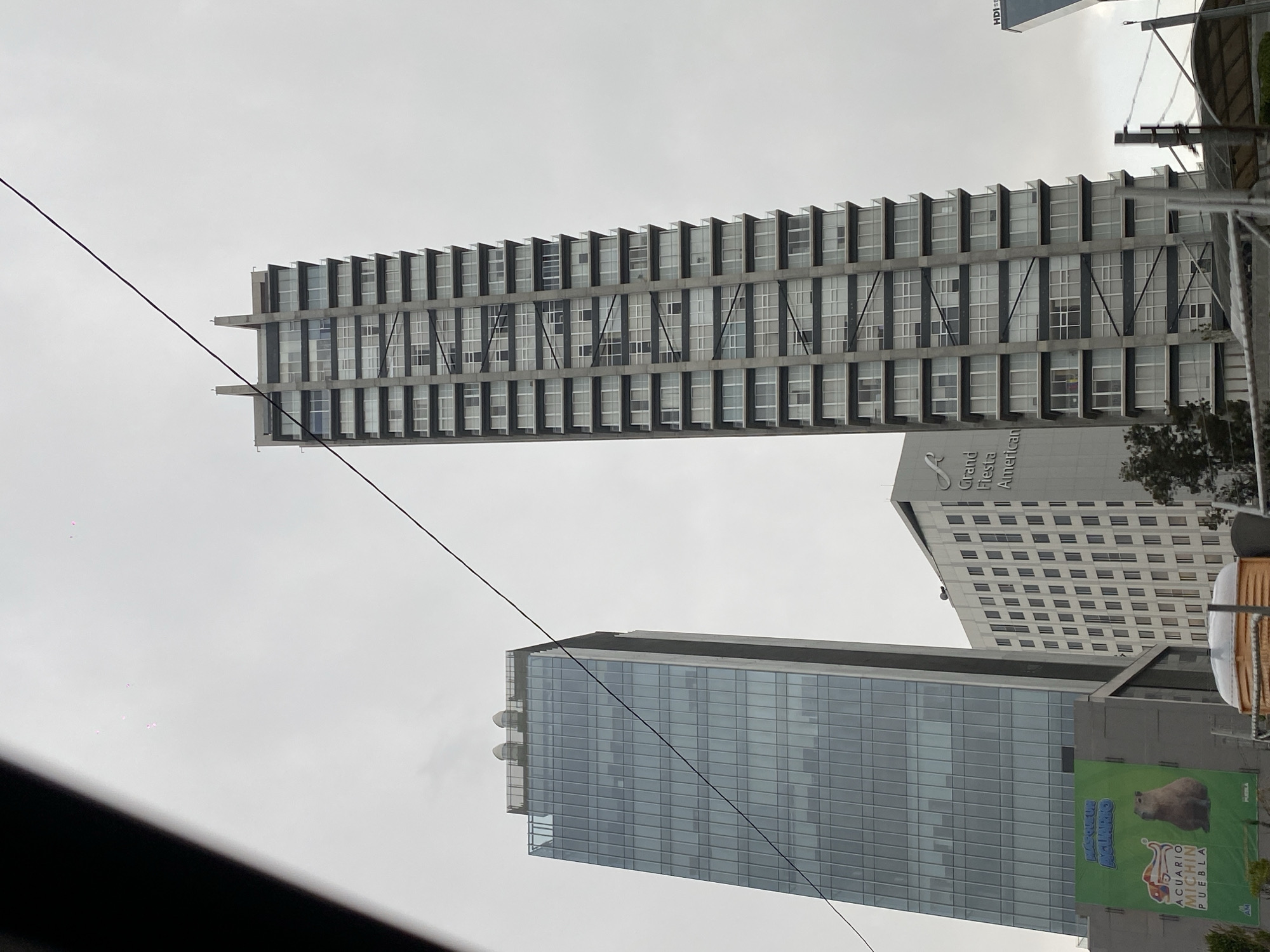 The image captures a cityscape featuring several modern buildings under an overcast sky. A tall, rectangular building with a distinctive façade of regularly spaced windows and concrete supports dominates the upper portion of the frame. Below it, another skyscraper, primarily glass-clad, rises, reflecting the ambient light. To the right, a building with "Grand Fiesta Americana" signage is visible. A large banner advertising "ACUARIO MICHIN PUEBLA" is attached to one of the buildings. A power line diagonally bisects the frame. The photograph appears to have been taken from inside a vehicle, indicated by the blurred section at the bottom of the frame. The city location remains unknown.
