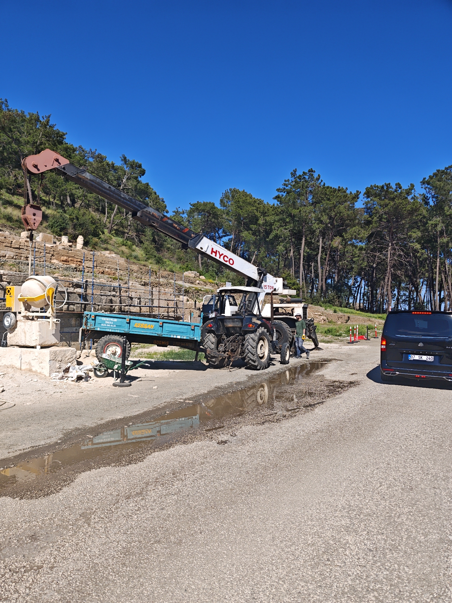 This media file depicts an active outdoor work site under a clear, vibrant blue sky. The scene is set on an unpaved, gravelly road with a significant puddle reflecting the surrounding environment.
In the center of the image, a black tractor is prominent, equipped with a large crane attachment. The crane's boom extends towards the left, displaying the brand name "HYCO" and a "15 TON" weight capacity. The tractor is hitched to a teal-colored trailer. Visible text on the trailer includes "COŞKUNLAR DORNA", a phone number "0505 066 61 61", "HESBAN", and "ÖDEMİŞ" with another phone number "02325444874". Adjacent to the tractor, a yellow concrete mixer sits on a stone base, with a green wheelbarrow positioned nearby.
In the background, behind the construction equipment, are ancient stone ruins, partially obscured by scaffolding, indicating an ongoing restoration or archaeological project. A dense forest of tall pine trees covers the hillside behind the ruins, extending across the upper background of the image.
On the right side of the road, a black Mercedes-Benz minivan, identified by the "MAYBACH" emblem on its rear, is parked. Its license plate clearly reads "07 SK 240". A man wearing a green shirt, light-colored trousers, and a cap is seen standing near the rear of the tractor, suggesting he is a worker on the site. Further along the road, orange and white traffic cones are visible.
The bright, clear sky and distinct shadows suggest it is a sunny day, likely late morning or early afternoon. The puddle on the road may indicate recent rain or water usage during construction. The overall scene points to a construction or restoration effort at an archaeological site, possibly in Kemer, Turkey.