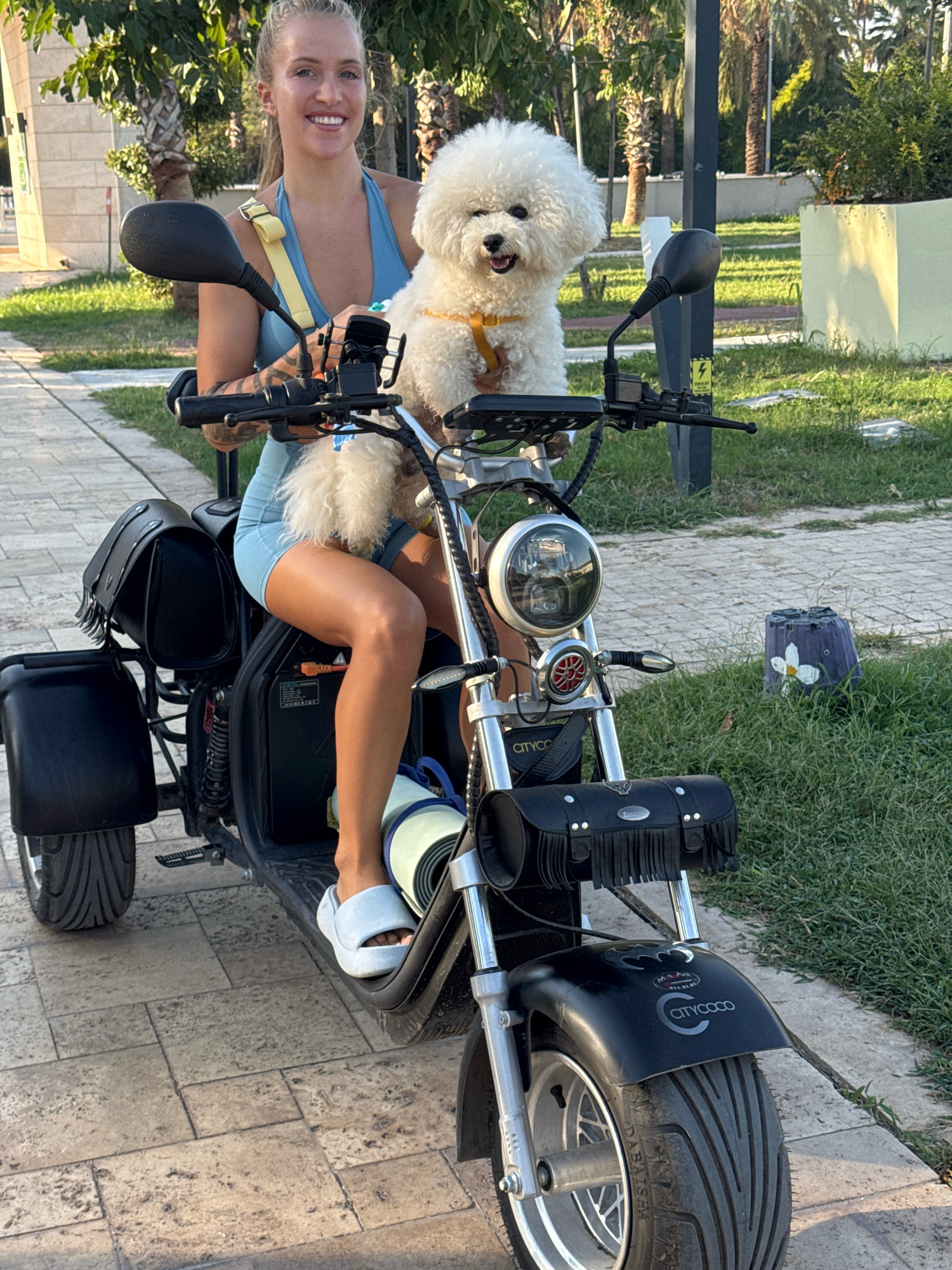 A blonde woman in a blue athletic outfit rides a black electric scooter with a Bichon Frise dog on her lap. The woman is smiling at the camera. The scooter has a large, round headlight and black saddlebags. The scene appears to be outdoors, possibly in a park or public area, with trees and green grass in the background. The pavement is made of stone tiles. There are some decorative items on the grass to the right of the scooter. The woman is wearing white slides. The dog is wearing a yellow harness.