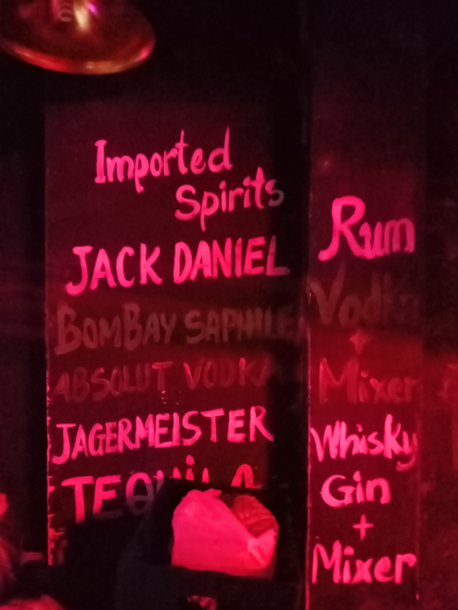 The image depicts a dimly lit interior of a bar or pub in Nha Trang, Vietnam, characterized by a dominant red and pink ambient light that creates a vibrant, neon-like atmosphere. The central feature is a vertical menu board, resembling a chalkboard, with handwritten text in a bright, glowing pink or red.
The left column of the menu is headed "Imported Spirits" and lists specific brands: "JACK DANIEL," "BOMBAY SAPPHIRE," "ABSOLUT VODKA," "JAGERMEISTER," and "TEQUILA." The right column presents generic drink categories and mixer options: "Rum," "Vodka," "Mixer" (appearing below Vodka), "Whisky," and "Gin + Mixer."
At the top of the frame, a blurry, reflective metallic object, possibly a light fixture or part of a ceiling fan, is visible, catching the red illumination. The bottom edge of the image shows indistinct reddish-pink shapes, which could be parts of hands or other items, partially obscured by the low light. The overall dimness and style of the lighting suggest the photograph was taken at night in a lively social setting. No identifiable individuals are clearly visible, but the menu implies customer presence.