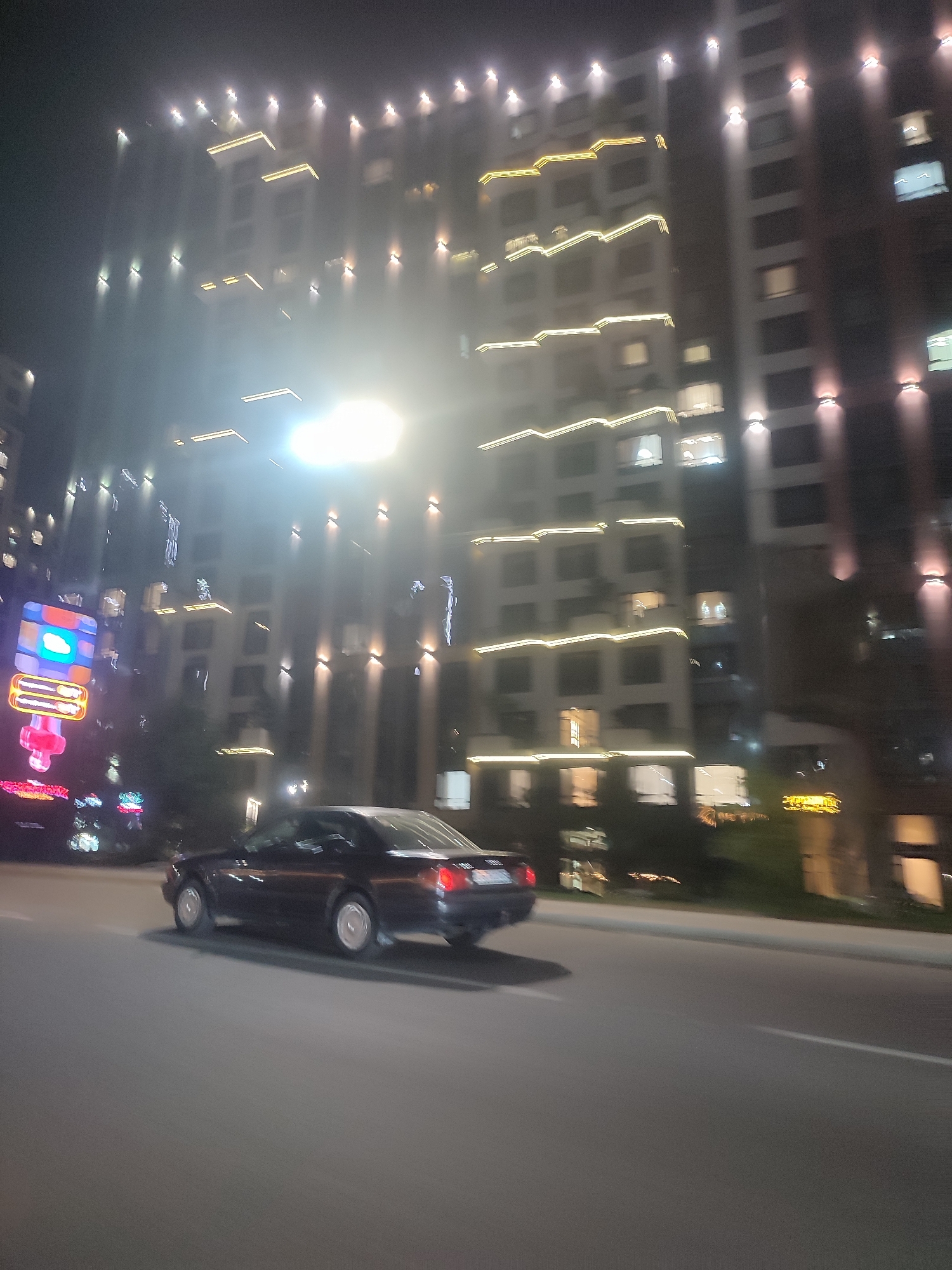 The image captures a nighttime street scene in Almaty, Kazakhstan, with a focus on a dark-colored sedan in motion. The car, possibly a coupe, is positioned in the right lane of a paved road and is captured with motion blur, indicating it is moving. Its taillights are illuminated red. In the background, a large, modern building is visible, adorned with multiple levels of decorative yellow and white lights that trace its architectural features. Smaller, individual lights are also mounted on the facade of the building at various intervals. To the left of the building, a brightly lit, multi-colored sign with geometric shapes and glowing elements is present. The overall environment appears to be an urban setting, characterized by the artificial lighting from the building and the sign, as well as the streetlights and the car's headlights, which are not clearly discernible due to the motion blur and night conditions. The road surface is dark asphalt with a white line marking the lane. The atmosphere is hazy, possibly due to fog or light pollution, which softens the edges of the distant structures and lights.
