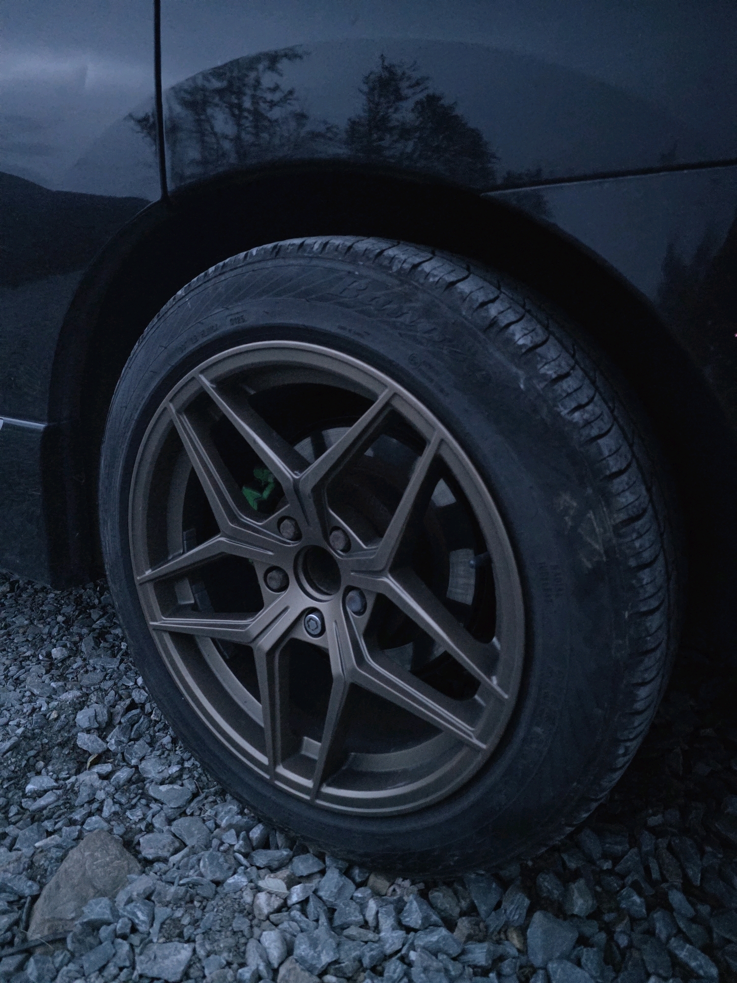 A close-up, low-angle shot of a car's wheel and tire is captured on a gravel surface in the city of Old Long Hill, Ireland. The car's dark body panels reflect trees and the sky, suggesting an overcast or twilight setting. The wheel features a bronze or gunmetal finish with a multi-spoke design. A bright green brake caliper is visible behind the spokes. The tire shows tread patterns and some markings. The gravel ground is composed of various sizes of grey stones. There are no people or visible actions in the image.