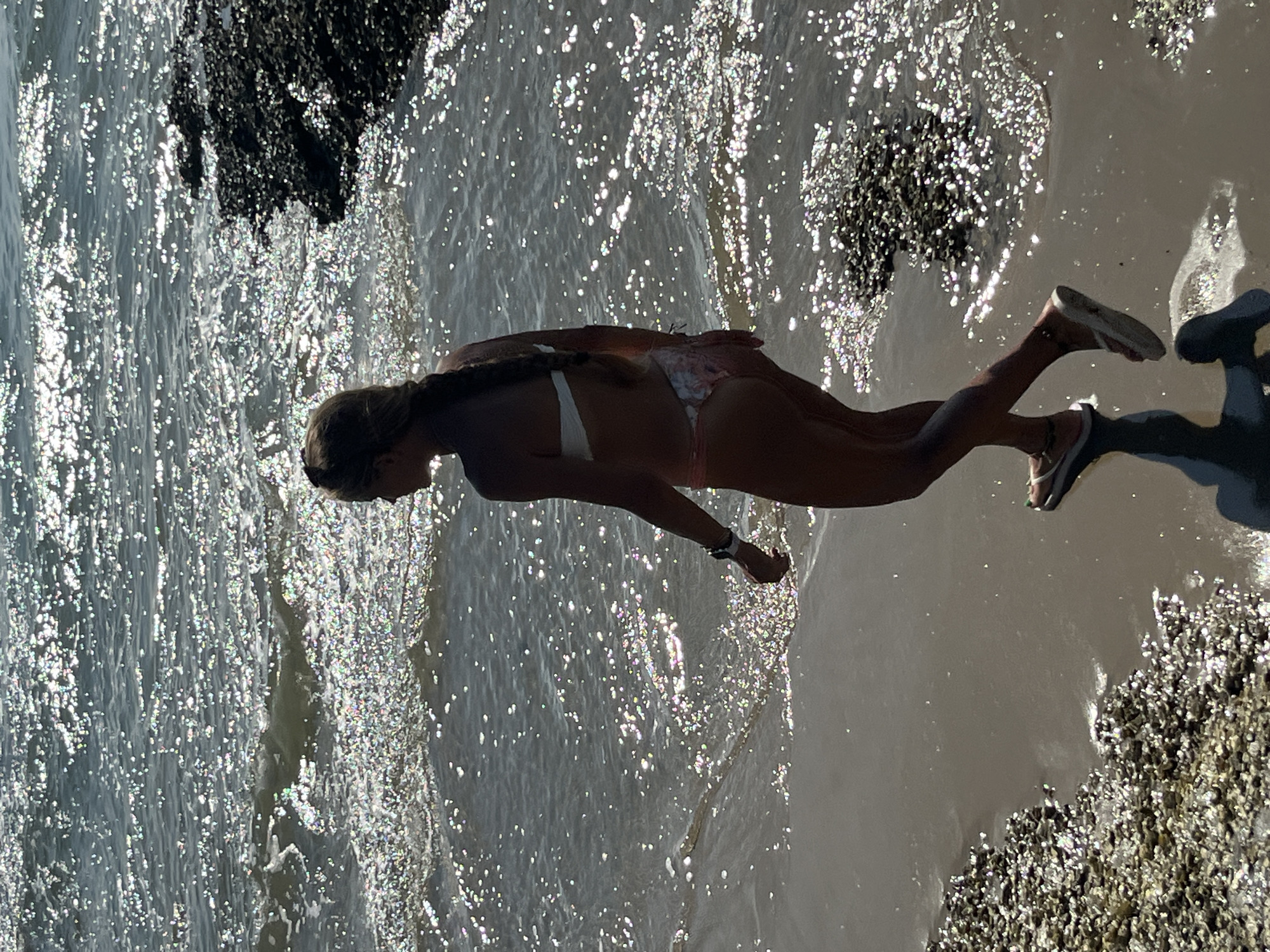 The image shows a woman in a bikini walking on a beach in Mũi Né, Vietnam. The sun is reflecting off the water, creating a shimmering effect. The woman is walking away from the camera, and her hair is in a braid. She is wearing a white bikini top and a patterned bikini bottom. The beach is covered in small shells and rocks. The water is clear and shallow. There is a rocky outcrop to the right of the image.