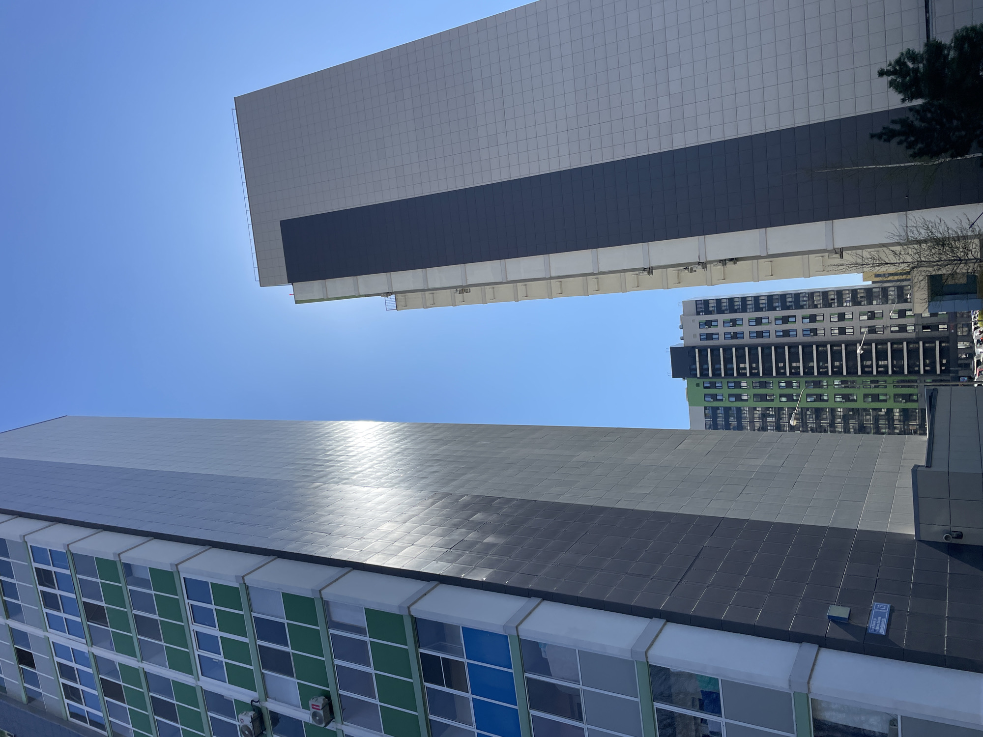 The image shows a low-angle view of modern buildings in Kazan, Russia. Two large, tiled buildings dominate the foreground, angled towards each other. The building at the bottom features a facade with square windows, some colored green and blue. The upper building is mostly gray, with a dark gray section. In the background, other high-rise buildings can be seen against a clear, blue sky. The bright sunlight suggests a daytime scene.