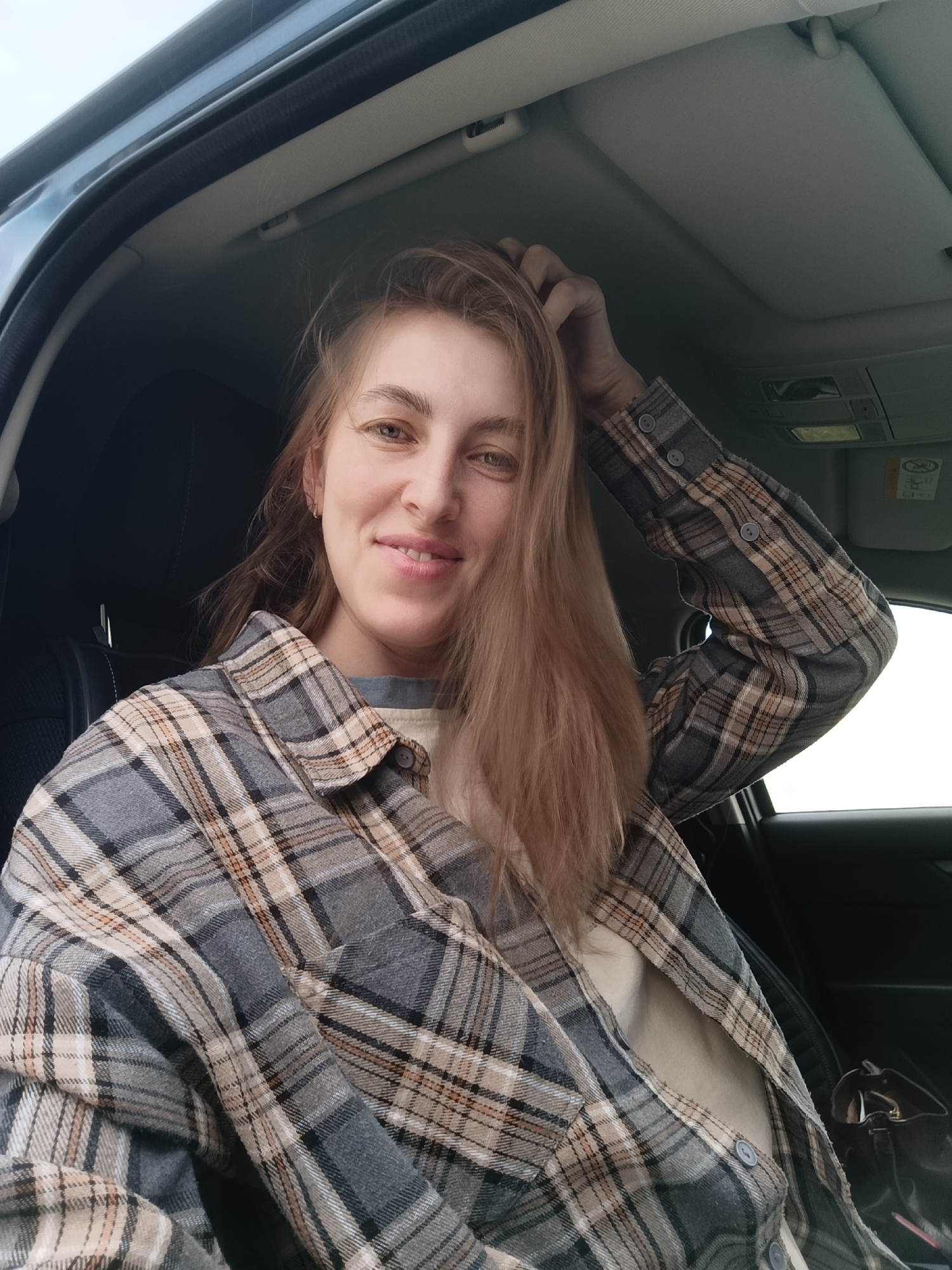 A woman is sitting in the driver's seat of a car. She is fair-skinned with light brown hair and is wearing a gray, brown, and white plaid shirt. Her right hand is raised, touching her hair. The car's interior is dark, with black seats. The car's roof and window are visible. The location is Chaikovsky, Russia.