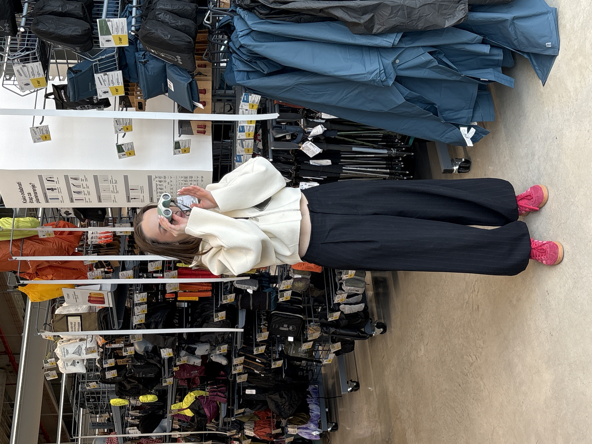 A young woman is depicted indoors, actively engaged in browsing a retail store specializing in outdoor equipment, likely a sports or hiking store. She is dressed in a cream-colored long-sleeved top, dark pinstriped wide-leg trousers, and vibrant pink sneakers. With a focused expression, she holds a pair of light green binoculars to her eyes, appearing to examine items on a distant shelf.
The store environment features extensive shelving and racks filled with various products. To her right, large stacks of folded blue jackets or raincoats are visible, with numerous walking or ski poles stored beneath them. To her left and behind her, shelves display an assortment of black bags, backpacks, and other camping or hiking accessories, including brightly colored orange and yellow fabric items. The floor is a light, textured concrete.
A prominent white display board in the mid-ground features text in Serbian: "Kako odabrati štap za planinarenje?" (How to choose a hiking pole?), accompanied by illustrative diagrams of different pole types. Several price tags are visible on various products, showing amounts like "2499" and "3499," likely in Serbian Dinars. The scene suggests a modern sporting goods store in Kragujevac, Serbia.