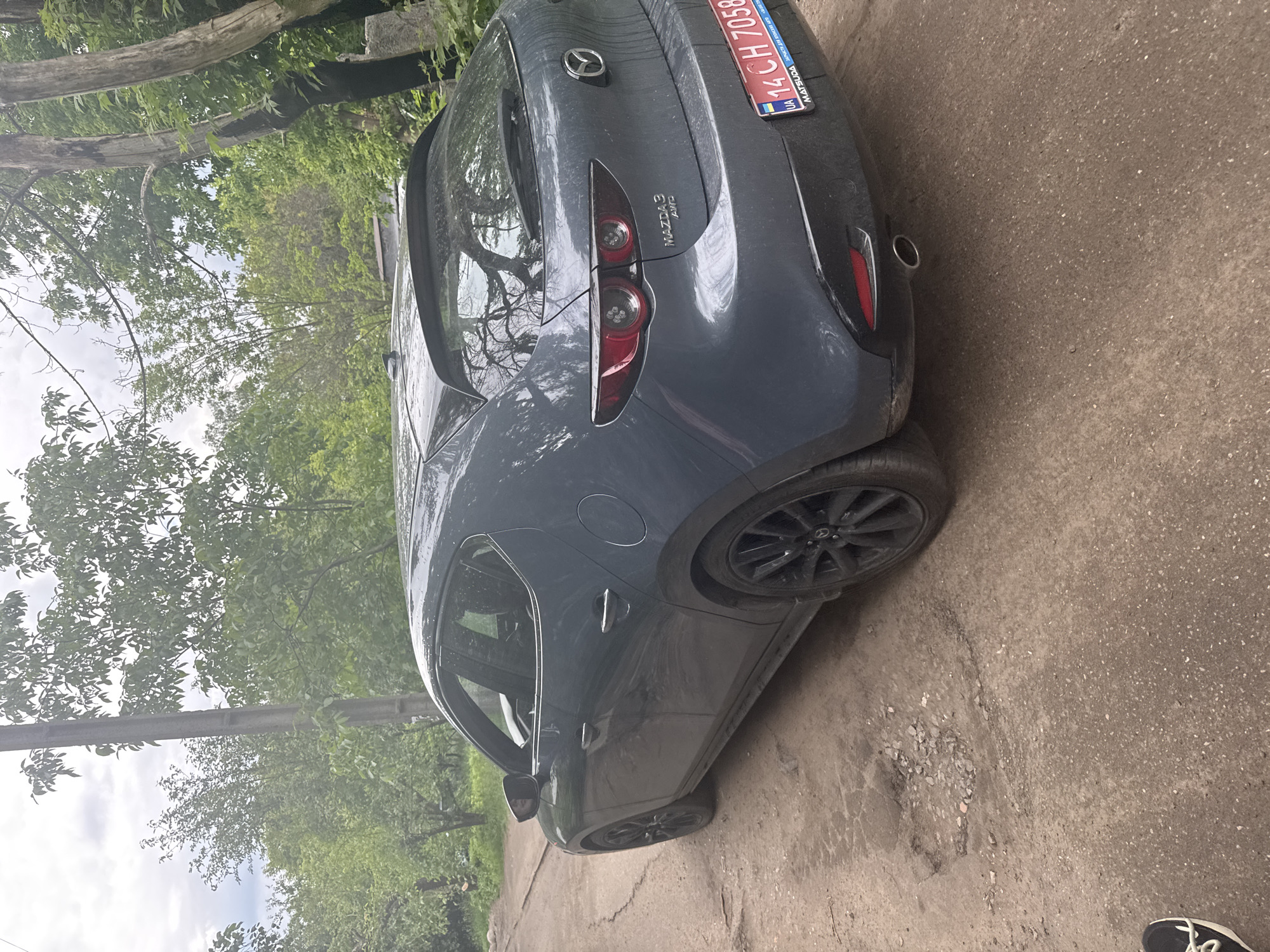 The image shows a dark gray Mazda 3 AWD hatchback parked on what appears to be a worn or unpaved road surface. The car is positioned at an angle, with its rear and left side visible. The vehicle appears somewhat dirty, possibly from recent travel or weather.
The rear of the car features distinctive red LED taillights. Below the Mazda emblem, the text "MAZDA 3 AWD" is clearly visible, indicating the model and drivetrain. A red and blue Ukrainian license plate, partially visible, displays the characters "14 CH 7058 BC" along with "UA" and "MATSUDA". A single exhaust pipe is visible on the right side of the rear bumper. The car is equipped with black multi-spoke wheels.
The setting is outdoors during the daytime. The sky is partially cloudy, and numerous green trees with lush foliage surround the area, suggesting a rural or suburban environment. Some tree branches extend over the car. The ground beneath the car is a rough, dark gray surface with visible cracks and scattered small stones, characteristic of an old asphalt or dirt road. In one of the cropped images, the tip of a person's black and white shoe is visible at the bottom right corner, suggesting someone is standing near the car.