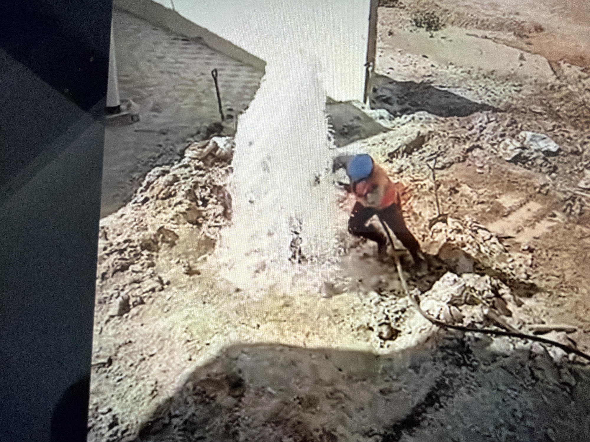 The image shows a construction worker wearing a blue hard hat and an orange vest operating a jackhammer in a muddy area. Water is spraying forcefully from a hole in the ground. The environment appears to be a construction site with dirt, rocks, and some debris. A shovel is visible in the background. The scene is likely related to construction or repair work, possibly involving underground utilities or water sources. The provided metadata does not contain information about the specific location in Orenburg, Russia.