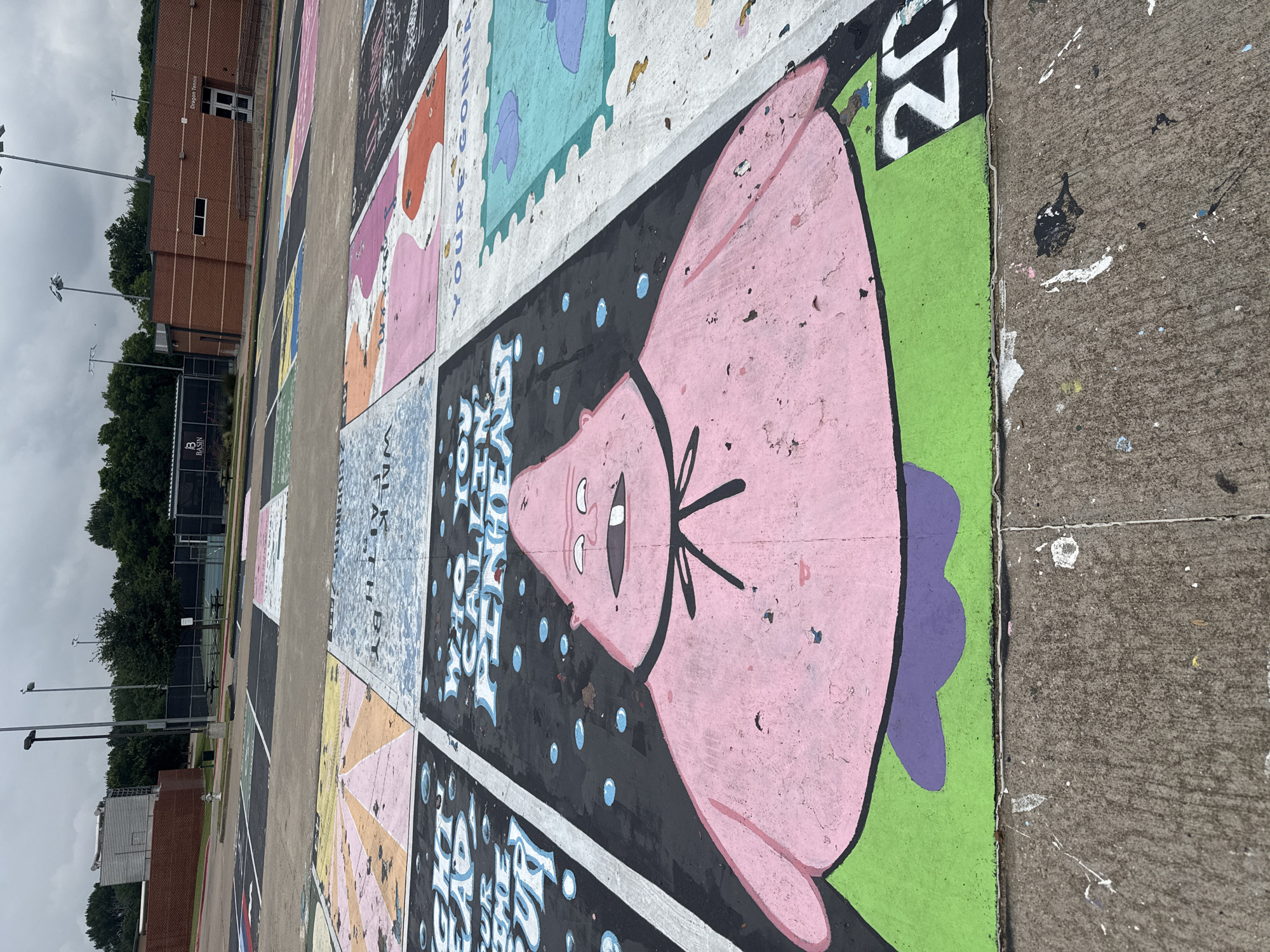 The image shows a section of a parking lot in Southlake, Texas, United States. The parking spaces are individually painted with different designs. One space features a large depiction of Patrick Star from Spongebob Squarepants, painted in pink, black, green and purple. Other spaces exhibit designs with stamps and written text. The asphalt in the parking lot has splattered paint. Beyond the parking spaces is a low brick building, likely associated with the parking area, and beyond that are trees and an overcast sky.