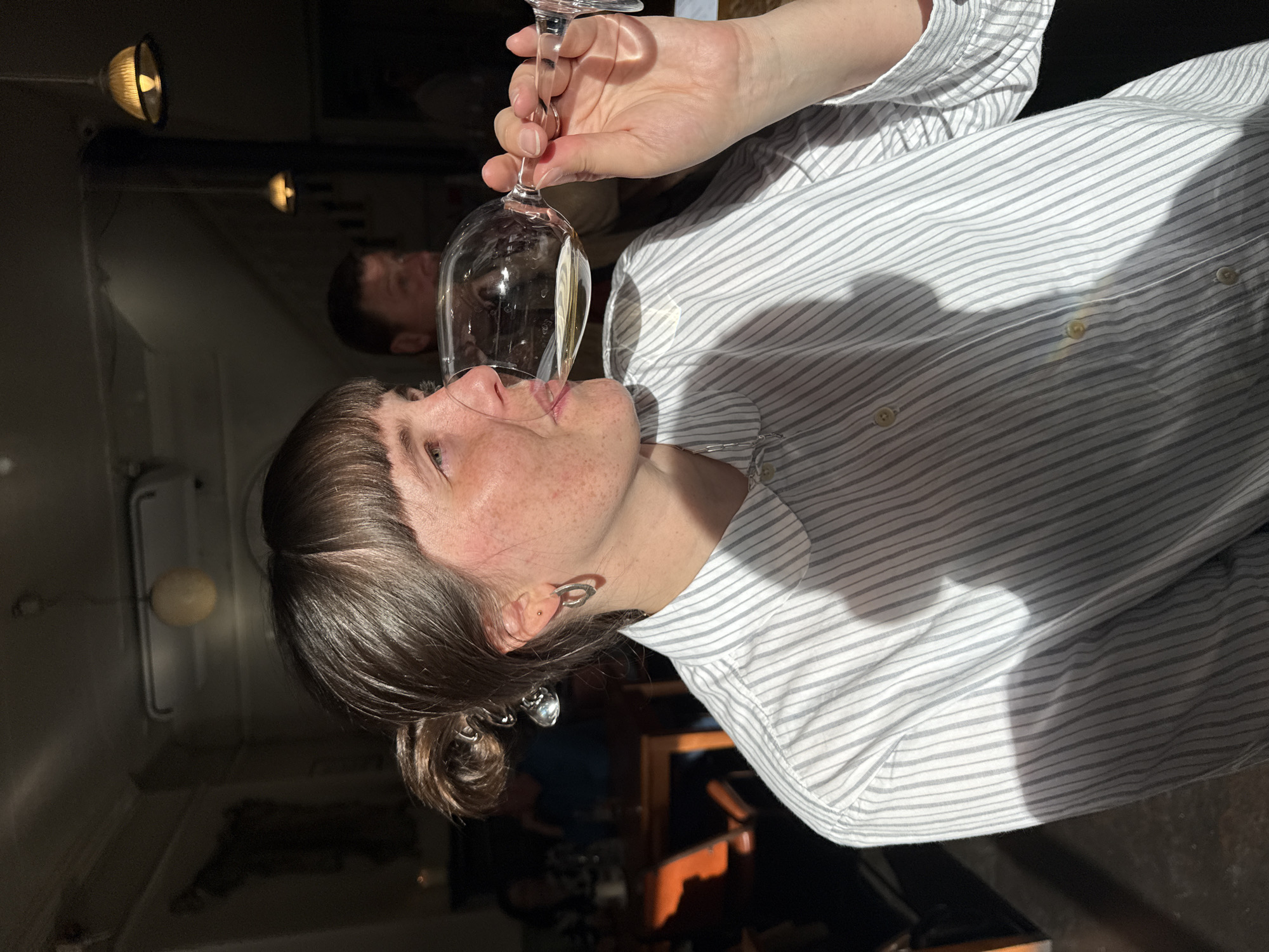 A person with brown hair in a bun, wearing a light gray and white striped shirt, is drinking from a wine glass filled with a light-colored liquid. Another person is partially visible in the background. The scene is indoors, possibly in a restaurant or bar. The location cannot be determined with the given information.