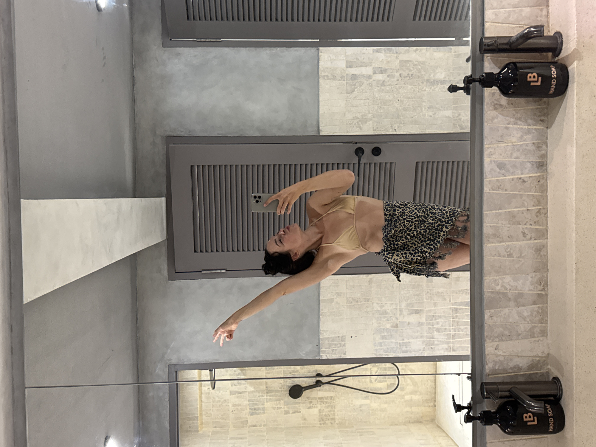 A woman in a bikini top and a leopard print sarong takes a mirror selfie in a modern bathroom. She is standing in front of a large mirror, with her left arm raised and bent, her hand forming a peace sign. Her right arm is holding a smartphone to take the picture. The bathroom features a tiled shower enclosure to the left, gray louvered doors behind her, and a double sink with soap dispensers and faucets in the foreground. The overall aesthetic is minimalist and chic, with concrete-effect walls and countertops. The location is a bathroom in North Kuta, Indonesia.