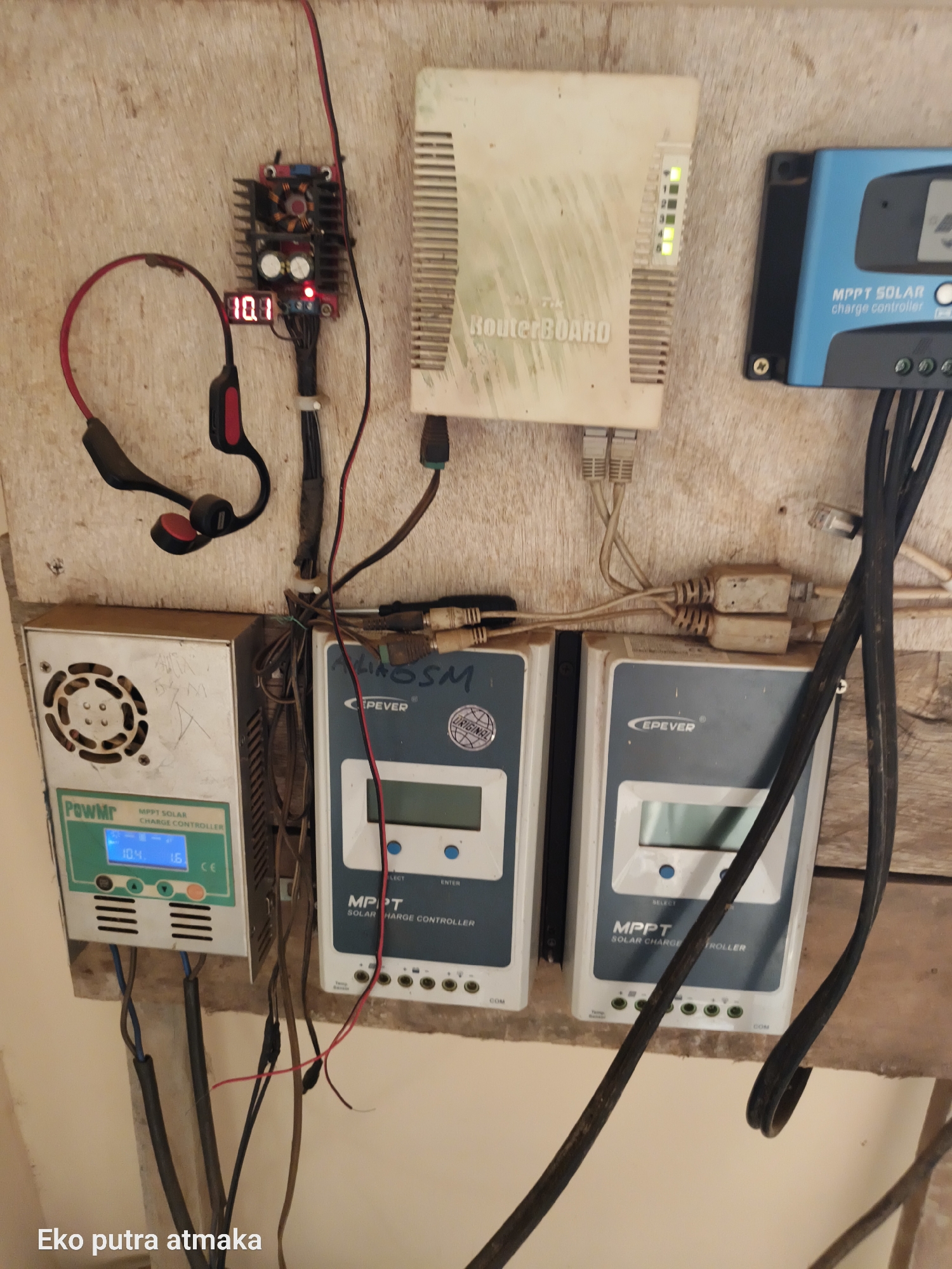 The image shows a wall-mounted setup of electronic devices, likely for a solar power system, in what appears to be an outdoor or semi-outdoor location. On the left is a "PowMr MPPT SOLAR CHARGE CONTROLLER" with a digital display showing "10.4" and "1.6". To its right is another "MPPT SOLAR CHARGE CONTROLLER" by "EPEVER". Above these are a "RouterBOARD" with several green indicator lights and a blue "MPPT SOLAR charge controller" which is partially obscured. A red digital display shows "10.1" near a black and red headset. The environment consists of a wooden board and various wires and cables connecting the devices. The text "Eko putra atmaka" is visible at the bottom left of the image. The location is identified as Pelanjau Jaya, Indonesia.