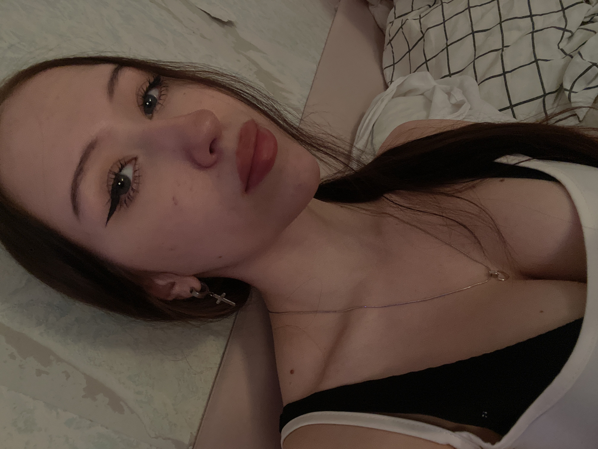 The media file shows a young woman lying on her side on a bed in Votkinsk, Russia. She has long dark brown hair and is wearing a white tank top over a black and white bra. She has dramatic winged eyeliner and red lipstick. A silver cross earring is visible on her left ear, and a thin silver necklace with a small pendant rests on her décolletage. The bedding behind her is white with a black checkered pattern. The lighting suggests it is nighttime or the room is dimly lit. She appears to be posing for the camera, with a relaxed expression.