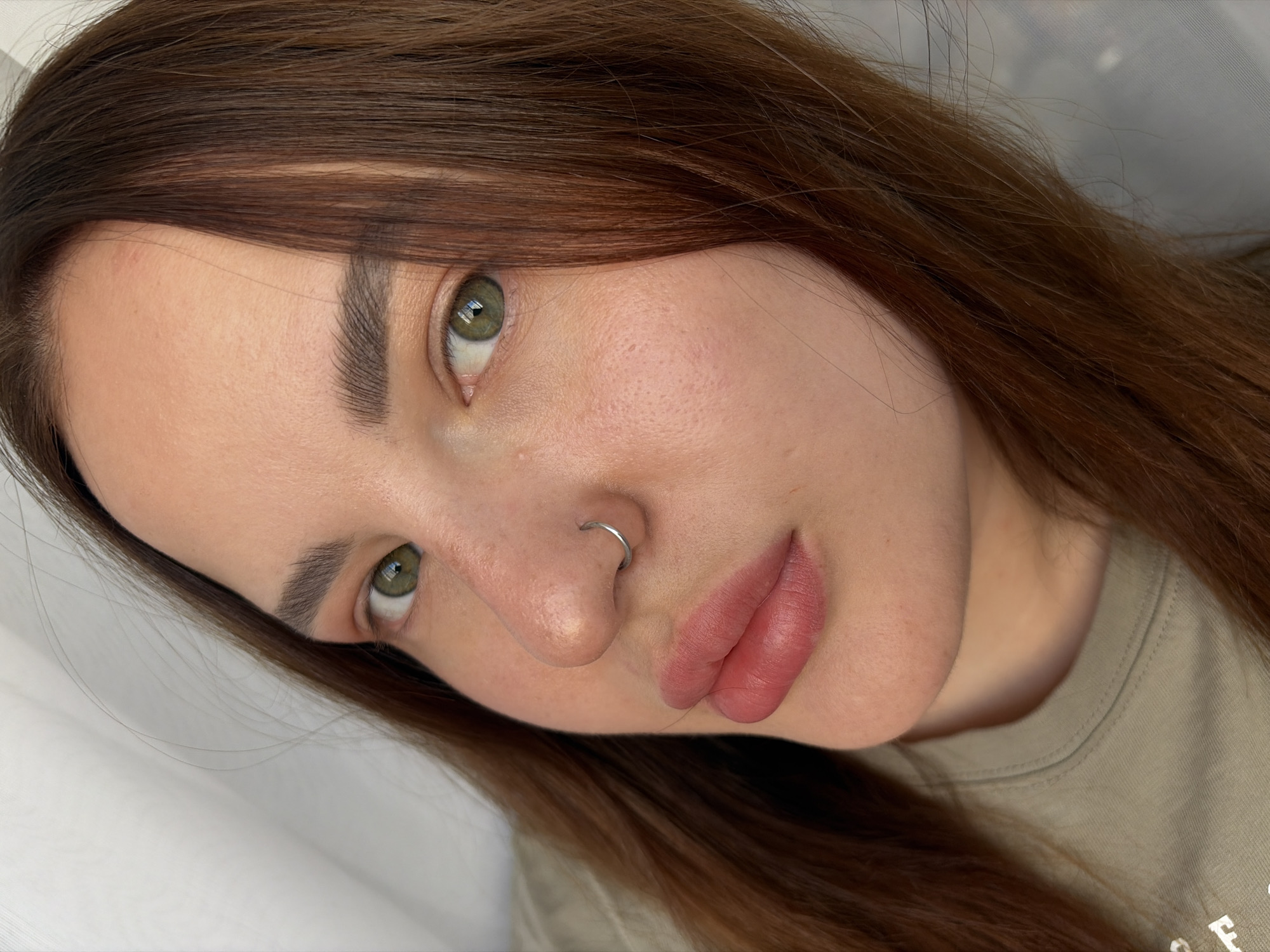 A close-up photograph captures a fair-skinned woman with green eyes, a nose ring, and brown hair. Her lips appear to have a rosy tint, possibly from lipstick or lip blushing. The woman is wearing a beige t-shirt. The background appears to be a white curtain.