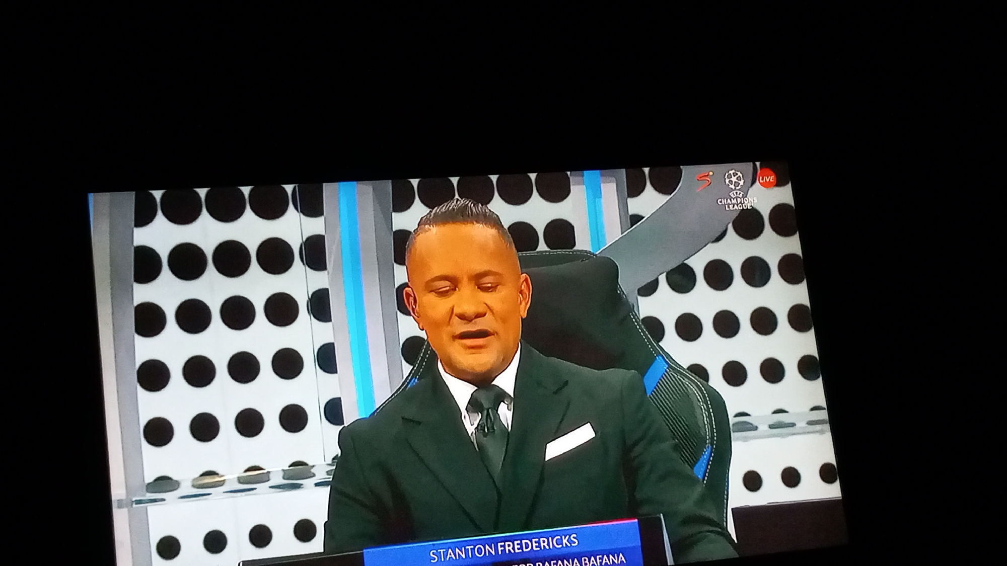 The image captures a man identified as Stanton Fredericks, likely a sports commentator or analyst, during a live broadcast. He is seated in what appears to be a broadcast studio, characterized by a striking background of regularly spaced black dots on a white grid, interspersed with vertical illuminated columns in blue and grey. The man wears a dark suit, a white shirt, and a dark tie, with a white pocket square. He has a short, styled haircut and his skin appears distinctly bronzed, possibly due to studio lighting or makeup. His mouth is slightly open, suggesting he is in the middle of speaking or listening intently, and his eyes are partly closed. He is seated in a modern black chair with blue accents, resembling a gaming chair.
In the upper right corner of the screen, a "LIVE" indicator is visible, along with the "UEFA Champions League" logo and a partial "SuperSport" (SSTV) logo, confirming the context of the broadcast. A lower third graphic at the bottom of the screen displays his name, "STANTON FREDERICKS," and partially visible text, "R BAFANA BAFANA," which refers to the South African national football team, indicating his expertise or connection to it. The overall setting is professional and modern, typical of a television sports studio. There are no direct visual cues in the image to confirm the provided location context of Nkpor, Nigeria.