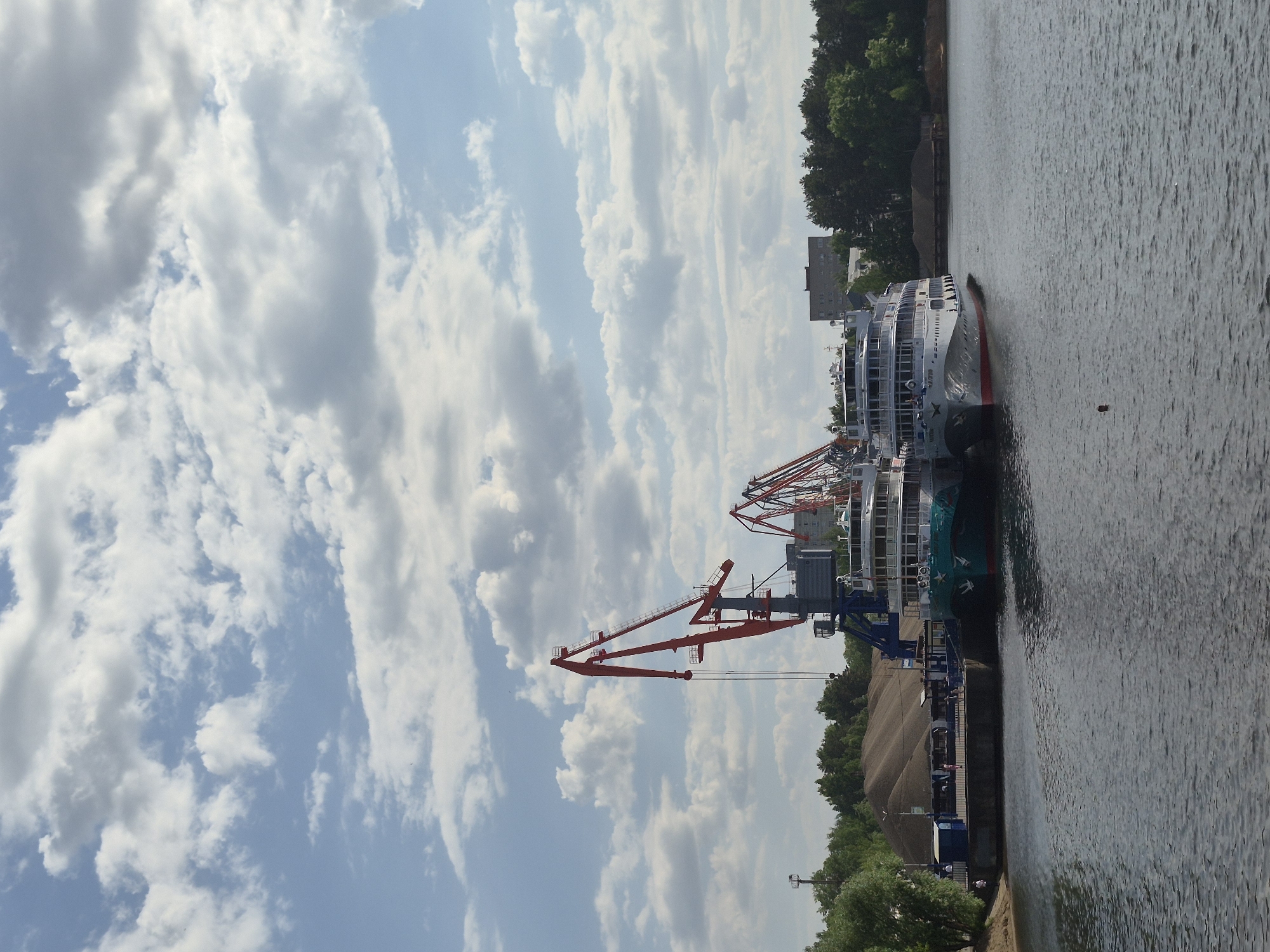 Here is a detailed description of the image:
The image captures a daytime scene at what appears to be a riverside port or docking area in the city of Unknown, Unknown. The view is oriented with the sky dominating the left side of the frame and the water on the right.
The sky is filled with a mix of fluffy white cumulus clouds scattered across a blue expanse. Several cranes, some in a striking red color, stand near the edge of the water, indicating cargo handling or shipyard activities.
The river is a dark, rippled surface, and docked alongside the pier is a large passenger ship, multiple decks visible. The ship is white with red accents and possibly green markings, and appears to be a river cruise or passenger vessel.
Along the riverbank, there are trees and some indistinct structures, including what looks like a pile of materials. The edge of the riverbank is lined with trees and some man-made structures. The perspective suggests the photo was taken from an elevated or slightly distant position. There are no people visible.