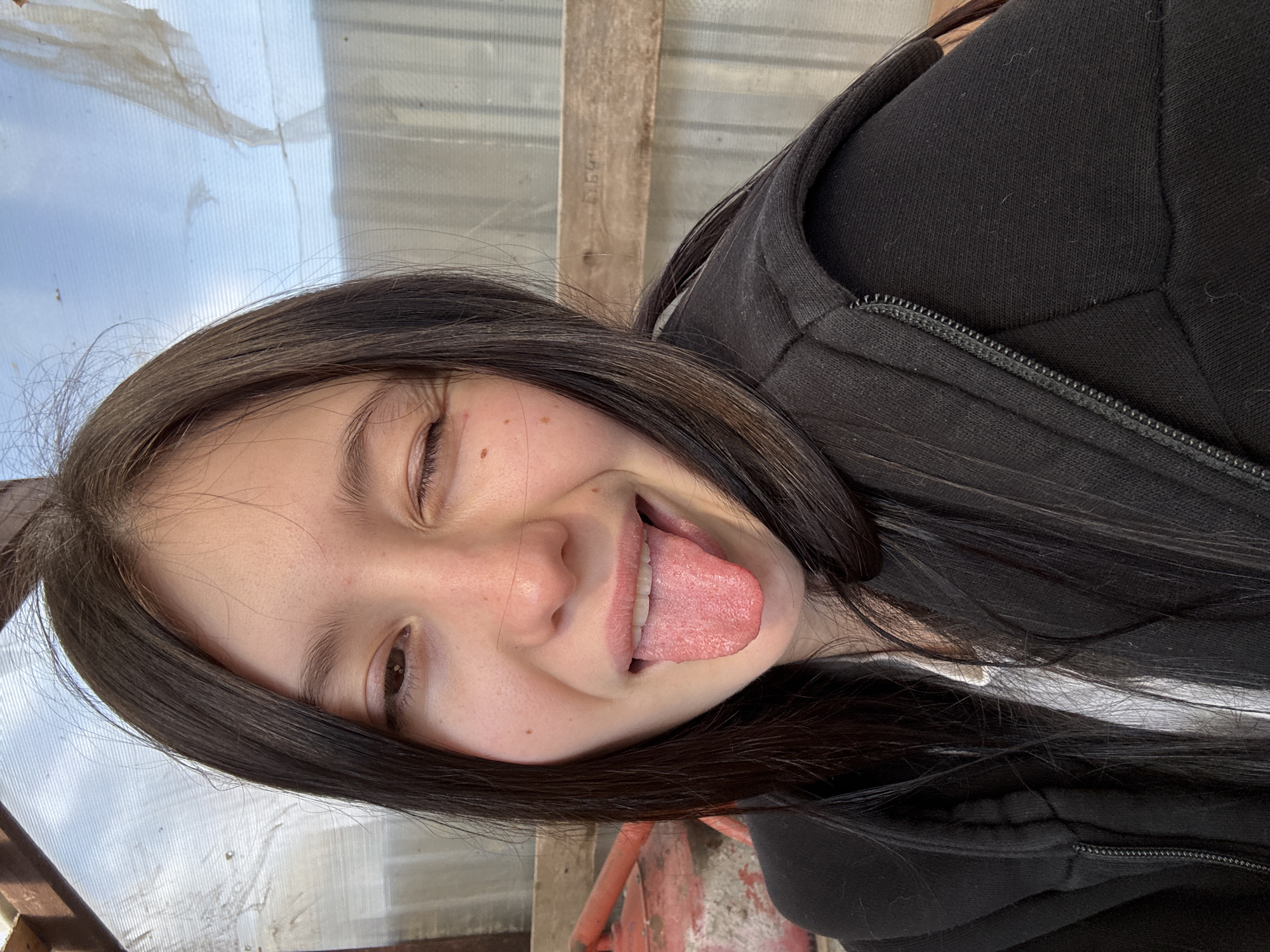 A young woman with dark hair, wearing a black zip-up hoodie over a white shirt, is making a silly face with her tongue sticking out and one eye squinted. The background appears to be a weathered structure with translucent panels, possibly a greenhouse or shed. Based on the provided location, it would be found in the town of Фоки, Russia. The foreground also reveals some wooden boards.