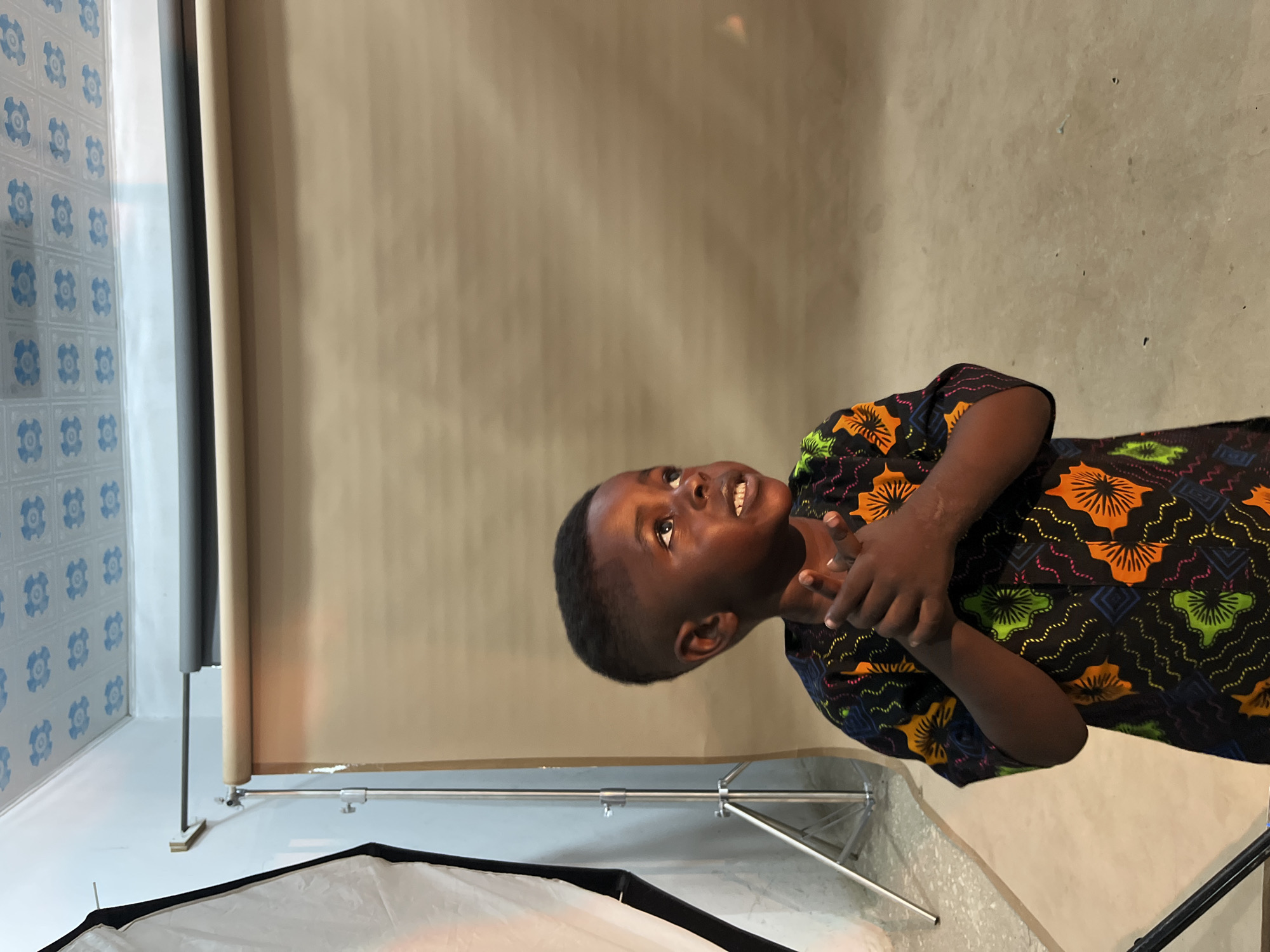 The image shows a young boy standing in what appears to be a makeshift photo studio. He is wearing a black shirt with a colorful African print featuring orange, green, and pink floral and geometric patterns. The boy is looking upwards and to the right, with his hands clasped together in front of his chest. His expression is one of curiosity or slight amusement.
The background consists of a large, textured brown paper backdrop, suggesting a professional or semi-professional photography setup. To the left of the boy, a lighting stand with a white diffusion panel is visible, as well as part of another background roller. Above, the ceiling is covered with white tiles featuring a blue, repeating floral pattern. The floor appears to be concrete. The overall impression is of an indoor setting, possibly a studio or a room adapted for photography. The location is specified as Gura Top, Nigeria.