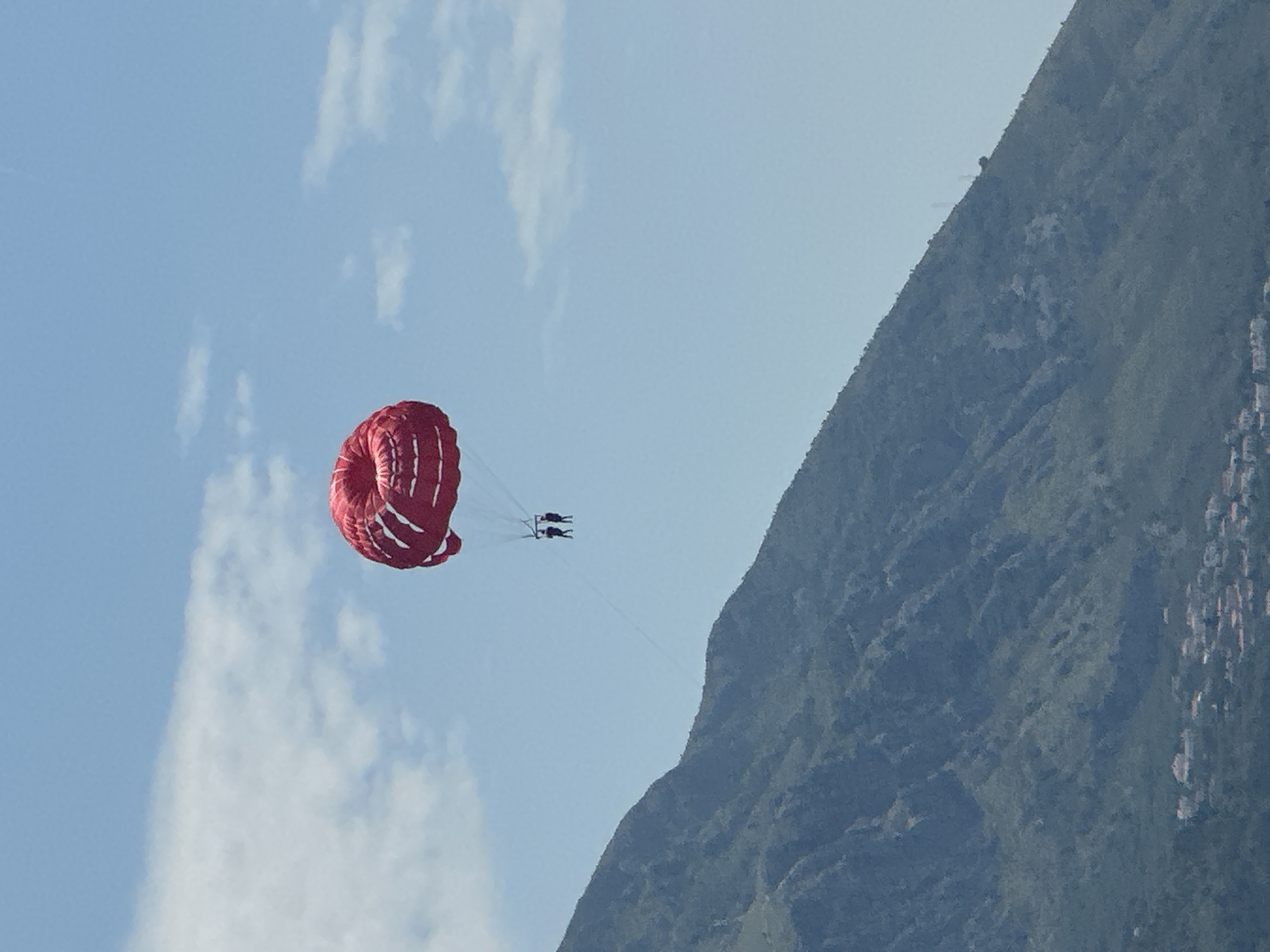 A red parachute with white accents is suspended in a clear blue sky, with scattered white clouds. Two individuals are attached to the parachute and appear to be parasailing. Below the parachute, a large, lush green mountain dominates the lower half of the image. The base of the mountain slopes down towards a distant, coastal town with red-tiled roofs, which is likely Boreti, Montenegro. The scene suggests a recreational activity taking place in a scenic, mountainous coastal region.