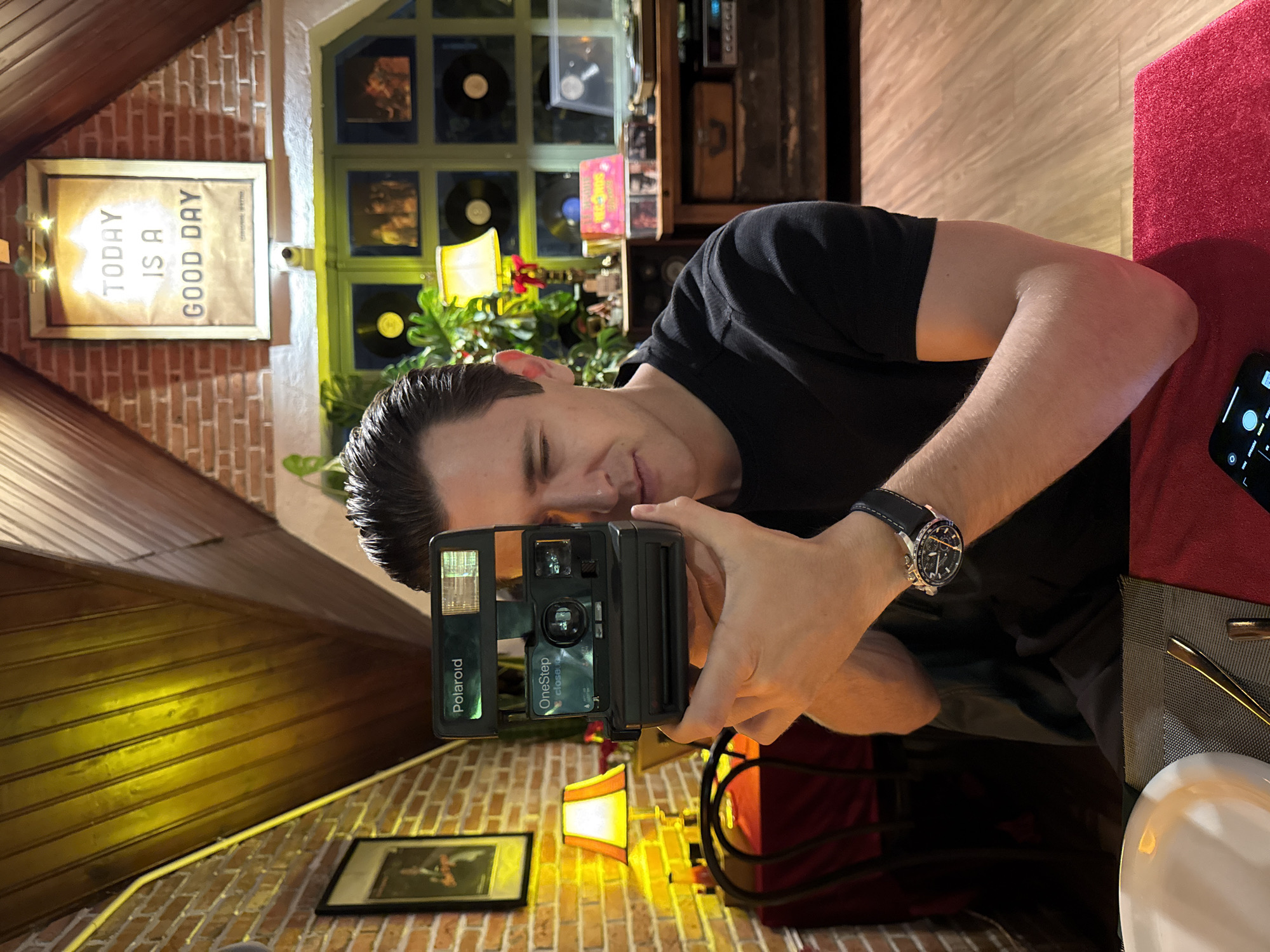 A man sits at a table inside a restaurant, holding a Polaroid OneStep CloseUp instant camera and looking into the viewfinder as if taking a picture. He is wearing a black t-shirt and a wristwatch. The restaurant has brick walls and wood-paneled ceilings. Decorations include framed records, a sign that reads "Today is a good day," and small lamps. On the table, there is a red tablecloth, cutlery, and a white plate. A phone is also visible on the table. The location may be in Lâm Viên - Đà Lạt, Vietnam.
