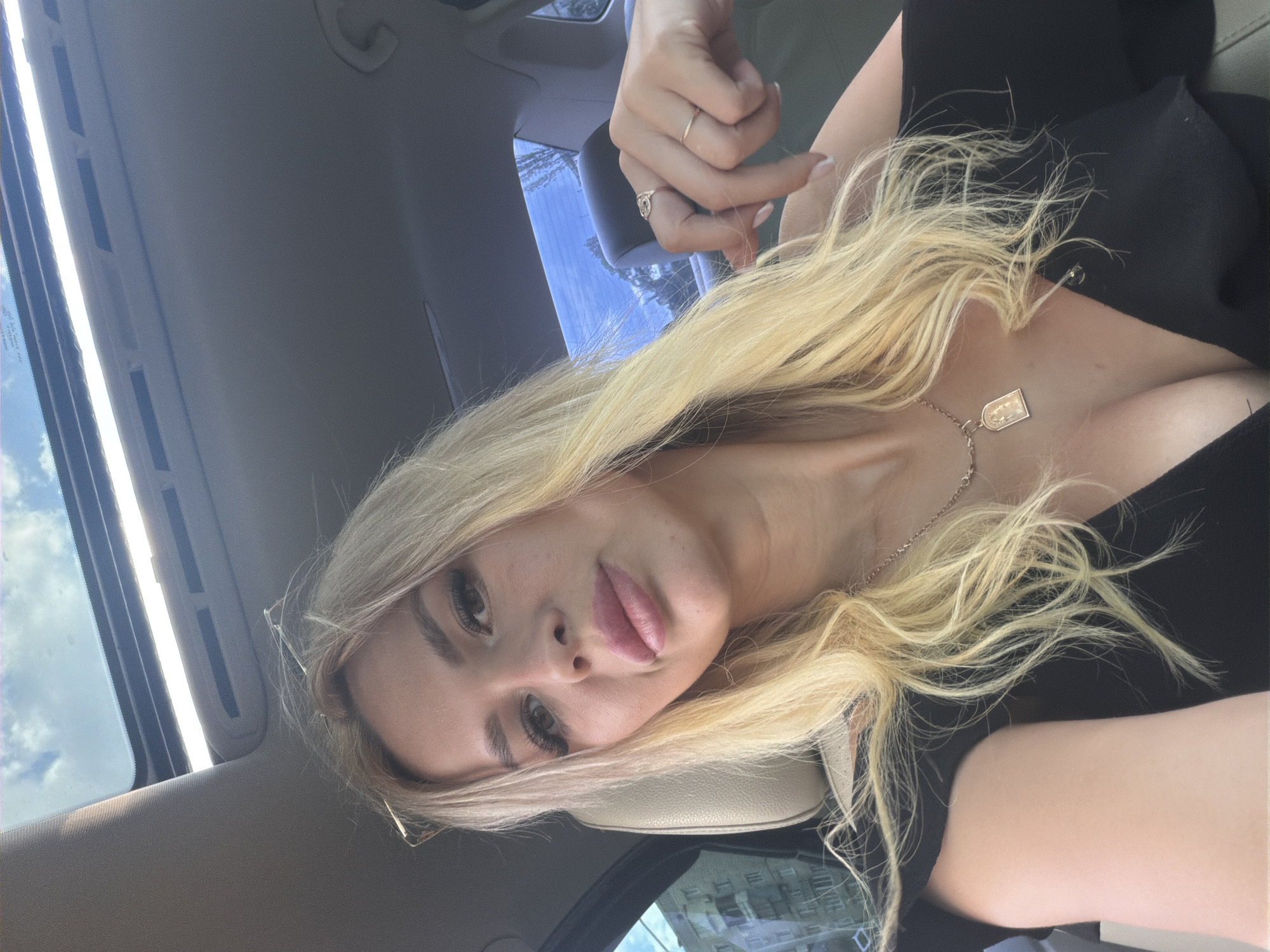 The image is a close-up, selfie-style shot of a young Caucasian woman with blonde hair and light skin. She is wearing a black top and a silver necklace with a rectangular pendant. Her hair is styled loosely, with some strands falling across her face and chest. She is wearing subtle makeup, including mascara and lipstick. She is wearing two rings on her fingers, one thin gold band and one with a rose-gold colored stone.
The woman is inside a car, as indicated by the car's interior visible in the foreground and background, including a beige car seat and part of the car's dashboard. Through the car's sunroof and window, a bright sky with some clouds is visible, suggesting it is daytime. The lighting indicates a sunny day. The overall mood is relaxed and casual.
The scene appears to be set in Kyiv, Ukraine. The image conveys a personal moment captured during a car journey, possibly on a sunny day. No visible text is present in the image.