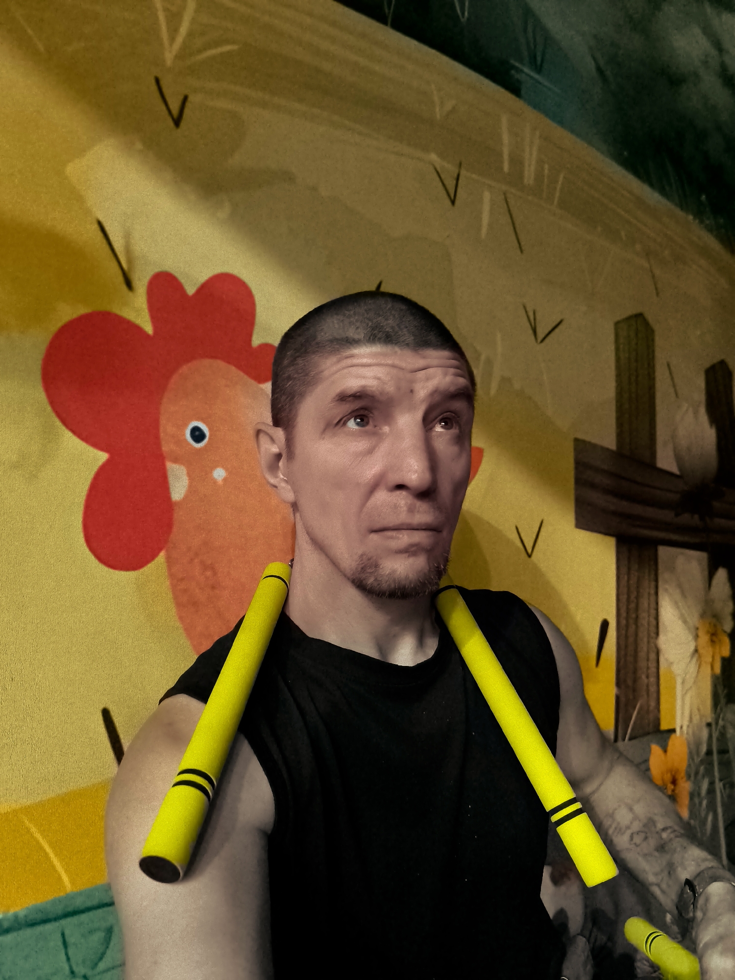 The image shows a man with a buzz cut and a goatee wearing a black tank top. He is holding two yellow cylindrical objects, each with two black bands, across his chest. The man is positioned in front of a mural or tapestry that features a cartoonish orange rooster with a red comb against a yellow background. To the right of the rooster, there is a depiction of a wooden cross and some flowers. The background appears to be indoors, possibly a room. There is no visible indication of the location being in Qarağandı city administration, Kazakhstan.