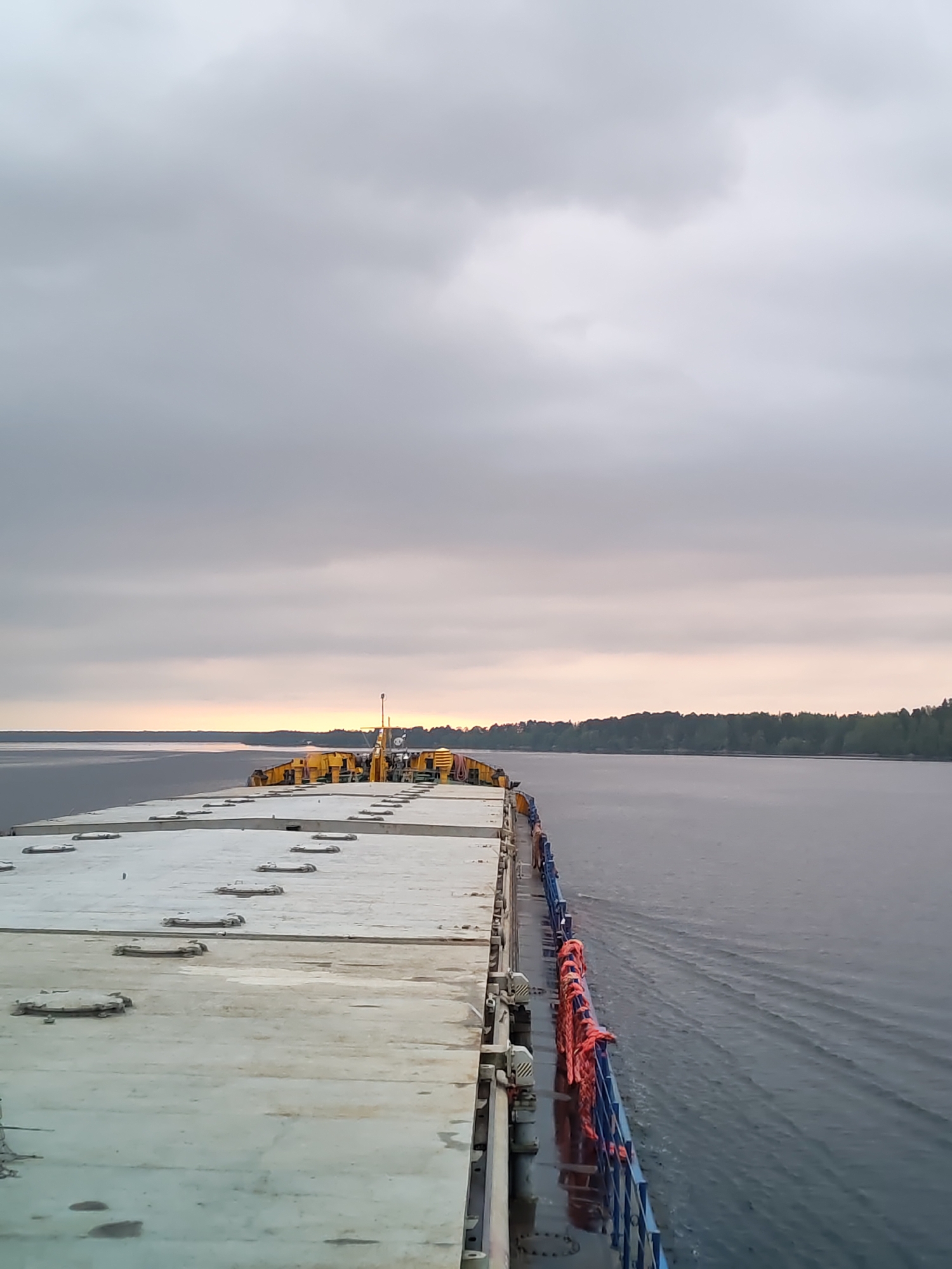 The image depicts the deck of a ship sailing on a body of water, possibly a wide river or a lake. The sky is overcast with gray clouds, and a hint of sunlight is visible near the horizon. The deck is light gray with a pattern of hatches or access points. The ship has a blue railing with orange life preservers or ropes attached. Trees are visible in the distance along the shoreline. The location is Unknown, Unknown.