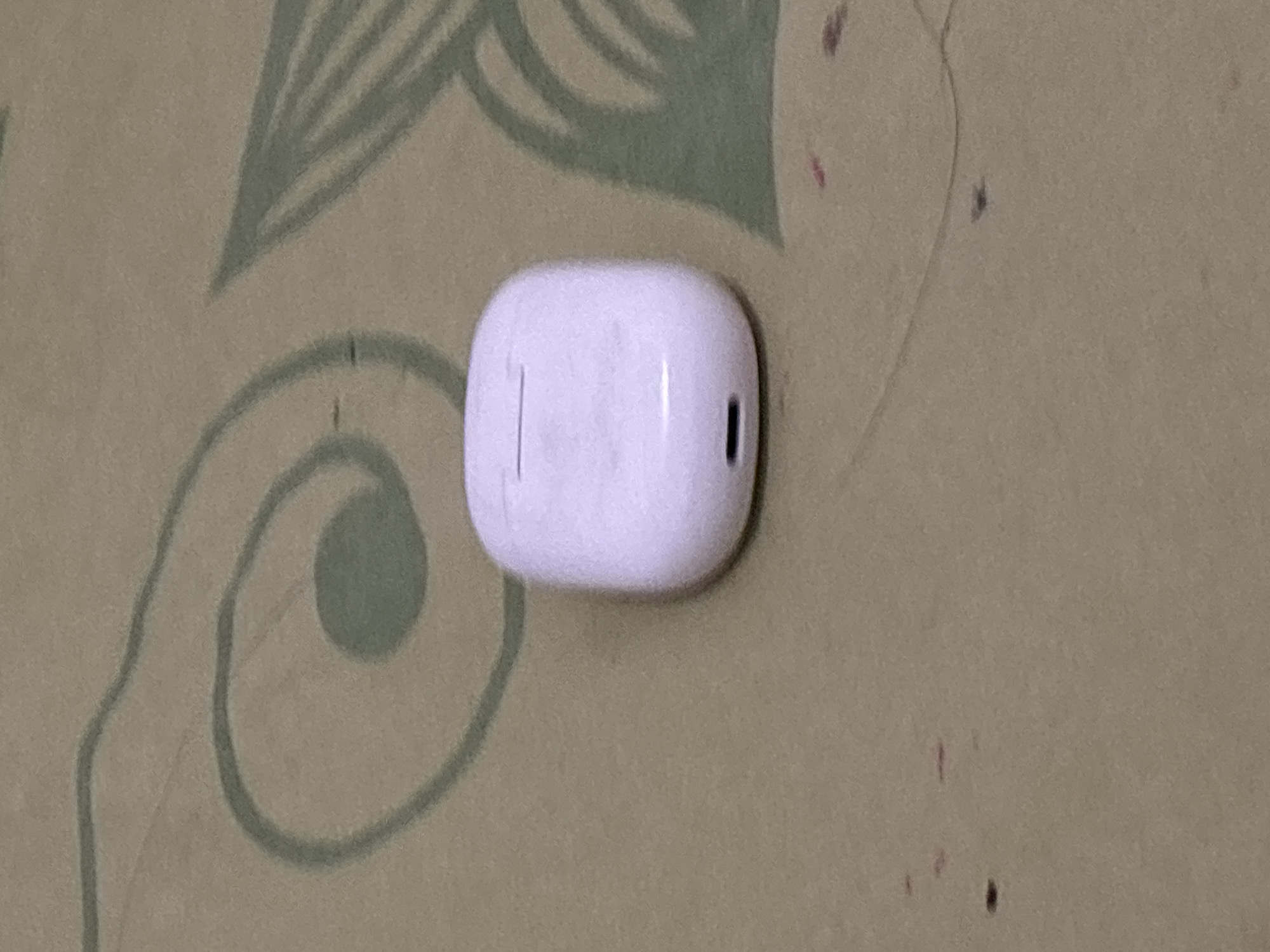 A white charging case for wireless earbuds is positioned centrally on a textured surface. The surface appears to be fabric, possibly a blanket or upholstery, with a faint, repeating pattern of green swirls and leaf-like shapes. Small, reddish-brown smudges are visible on the surface, scattered around the charging case. The charging case itself is oval-shaped with a smooth, matte finish. A thin, horizontal indentation is visible on the top of the case, indicating the seam where the lid opens. Near the bottom of the case, a small, dark rectangular slot is present, likely the charging port. The lighting in the image is dim, casting soft shadows and obscuring some finer details. The overall scene is domestic and appears to be an indoor setting. There is no identifiable information about the location being in North Jakarta, Indonesia.