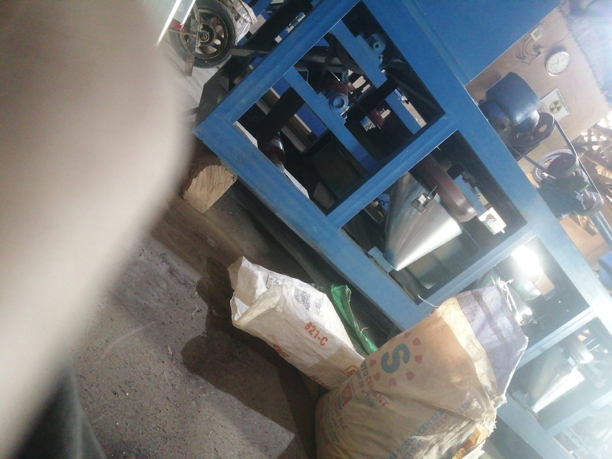 The image depicts an indoor scene, possibly a workshop or storage area, located in Jos, Nigeria. The main subject is a blue metal frame structure, likely a piece of machinery, with various components including pulleys and belts visible. To the left of the structure, there's a blurry foreground element, possibly a person or clothing, obscuring part of the scene. On the ground, there are two sacks, one white with red lettering and a QR code, and another larger, tan-colored sack with a prominent "S" logo. In the background, a clock is visible on the wall, and a circular symbol resembling a radiation warning is also present. Part of a wheel, possibly from a vehicle or equipment, is visible in the upper left corner. The environment appears somewhat utilitarian and potentially industrial.