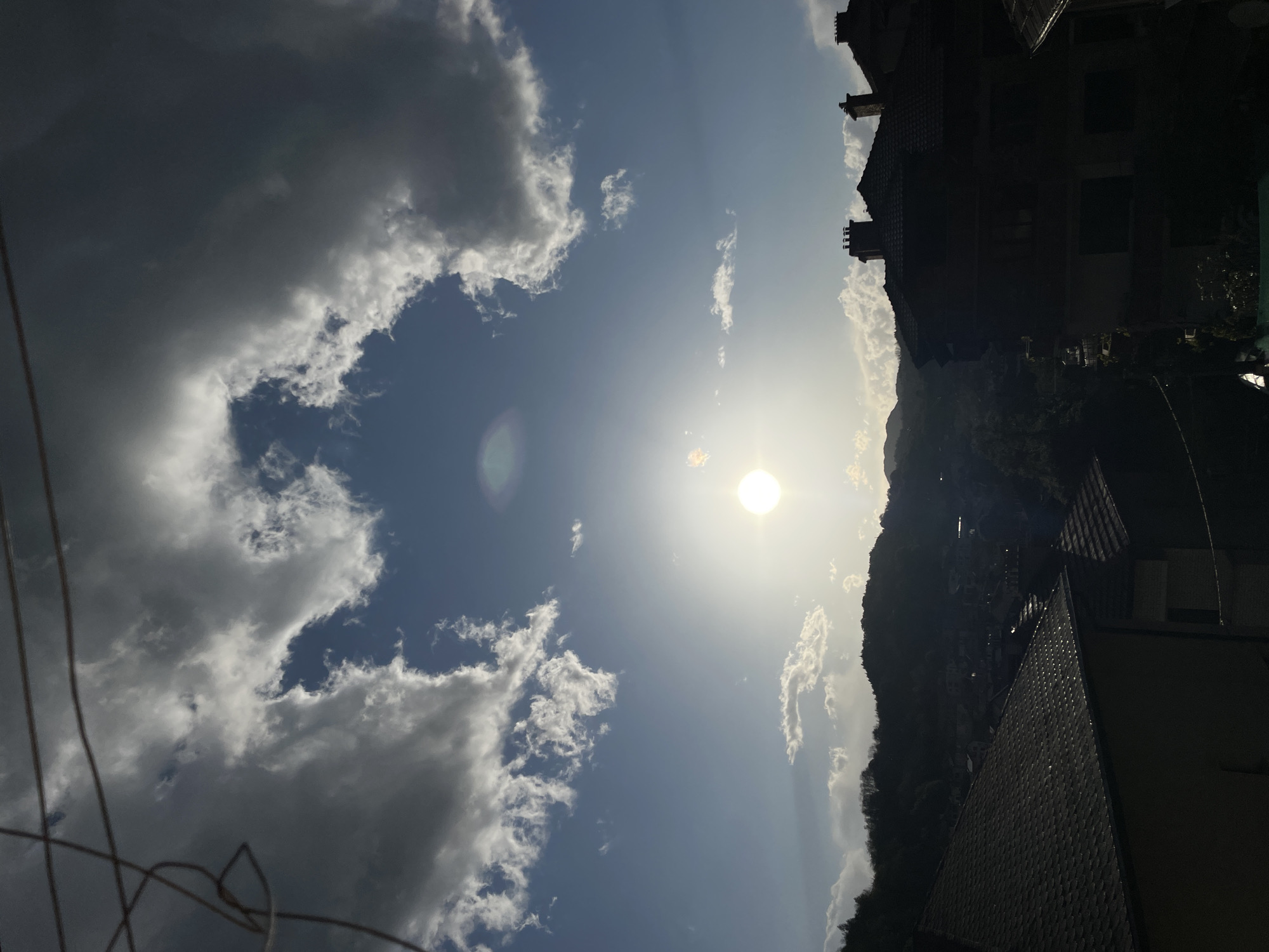 The image shows a view of the sky and rooftops in Zlatograd, Bulgaria. The sun is high in the sky, surrounded by thin clouds. There are larger, more defined clouds to the left. On the right side of the frame, there are several buildings, a mountain, and trees. A tangle of wires is visible in the lower-left corner. The image is taken from a slightly low angle, looking up towards the sky.