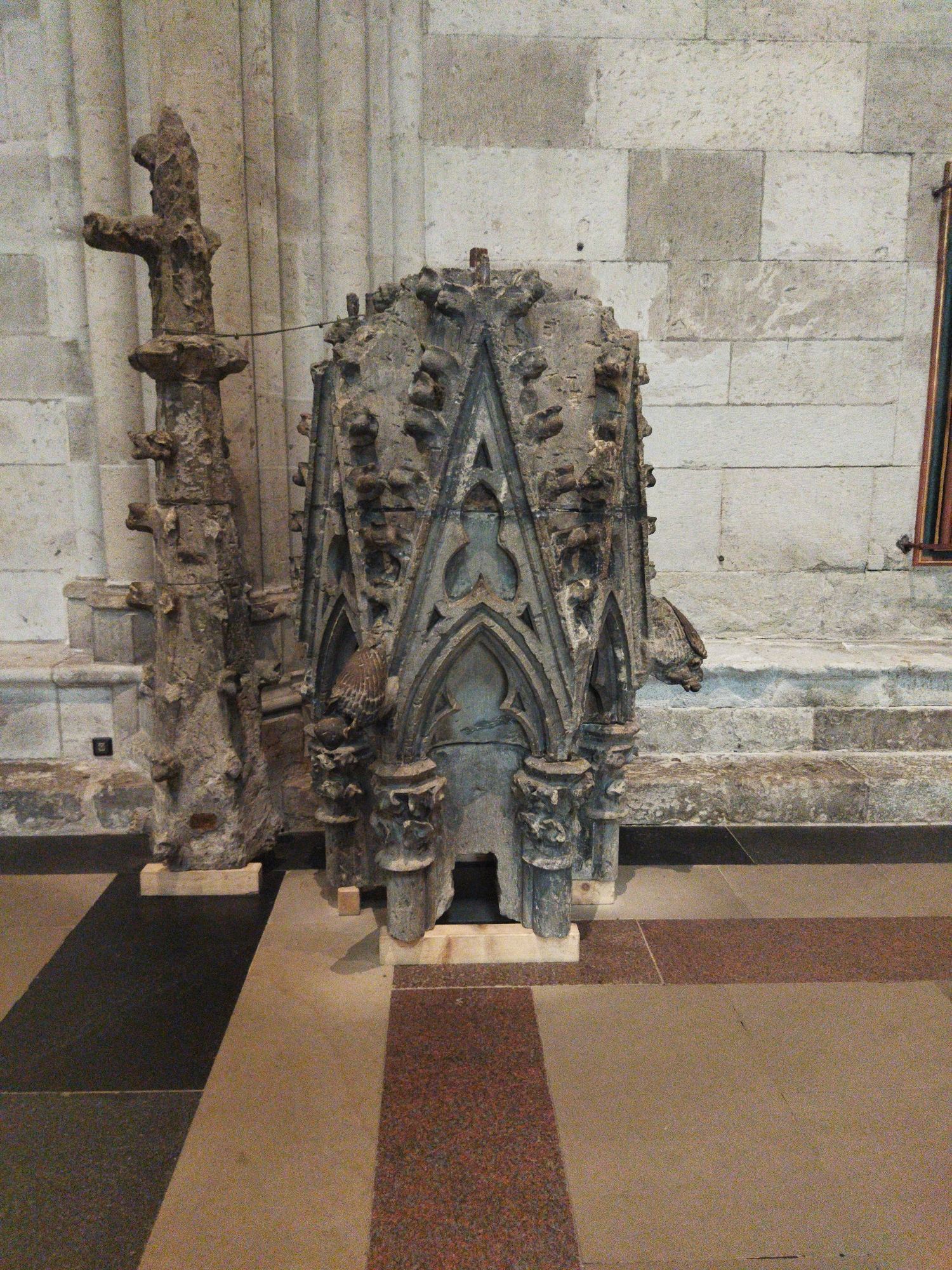 An ornate stone structure, resembling a miniature Gothic chapel or a large architectural fragment, is displayed indoors, possibly within a museum or a church in Monheim am Rhein, Germany. The structure is multifaceted with intricate carvings, pointed arches, and decorative elements. To the left of this main structure stands a tall, slender stone artifact, also exhibiting a rough, carved texture and a cross-like projection at its apex. Both artifacts are positioned on wooden supports atop a floor composed of large, square tiles arranged in a patterned design, with alternating sections of dark grey, beige, and reddish-brown granite. The background consists of a stone wall constructed from large, roughly cut blocks of light-colored stone, and vertical, fluted architectural elements that suggest the interior of a grand building. The lighting is ambient and even, casting no harsh shadows. There are no people or animals visible in the scene.