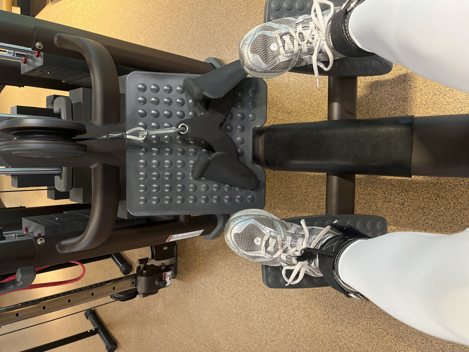 This overhead shot captures a person's lower body engaged with a gym machine within an indoor fitness facility, specifically located in North Kuta, Indonesia. The individual is wearing white athletic pants and silver/grey sneakers with white laces, which are secured to a textured, grey foot platform by black ankle straps.
The machine appears to be a cable-based strength training apparatus, possibly a leg press or a similar machine designed for leg exercises. A dark metal cable is visible, extending from a weight stack on the left (where dark grey weight plates are partially stacked) through a pulley system, and connecting to the foot platform via a metal carabiner. This setup indicates that resistance is applied through the cable. A padded, dark brown or black support bar is positioned horizontally between the person's feet. The machine's frame is robust, made of dark-colored metal.
The floor beneath the equipment is a light tan or brown color with a visible speckled texture, characteristic of rubberized gym flooring. The overall scene suggests an active moment during a workout, focusing on the interaction between the user and the exercise equipment. No specific time of day or emotions are discernible. A small white label with unreadable black text is present on the lower right side of the machine's frame.